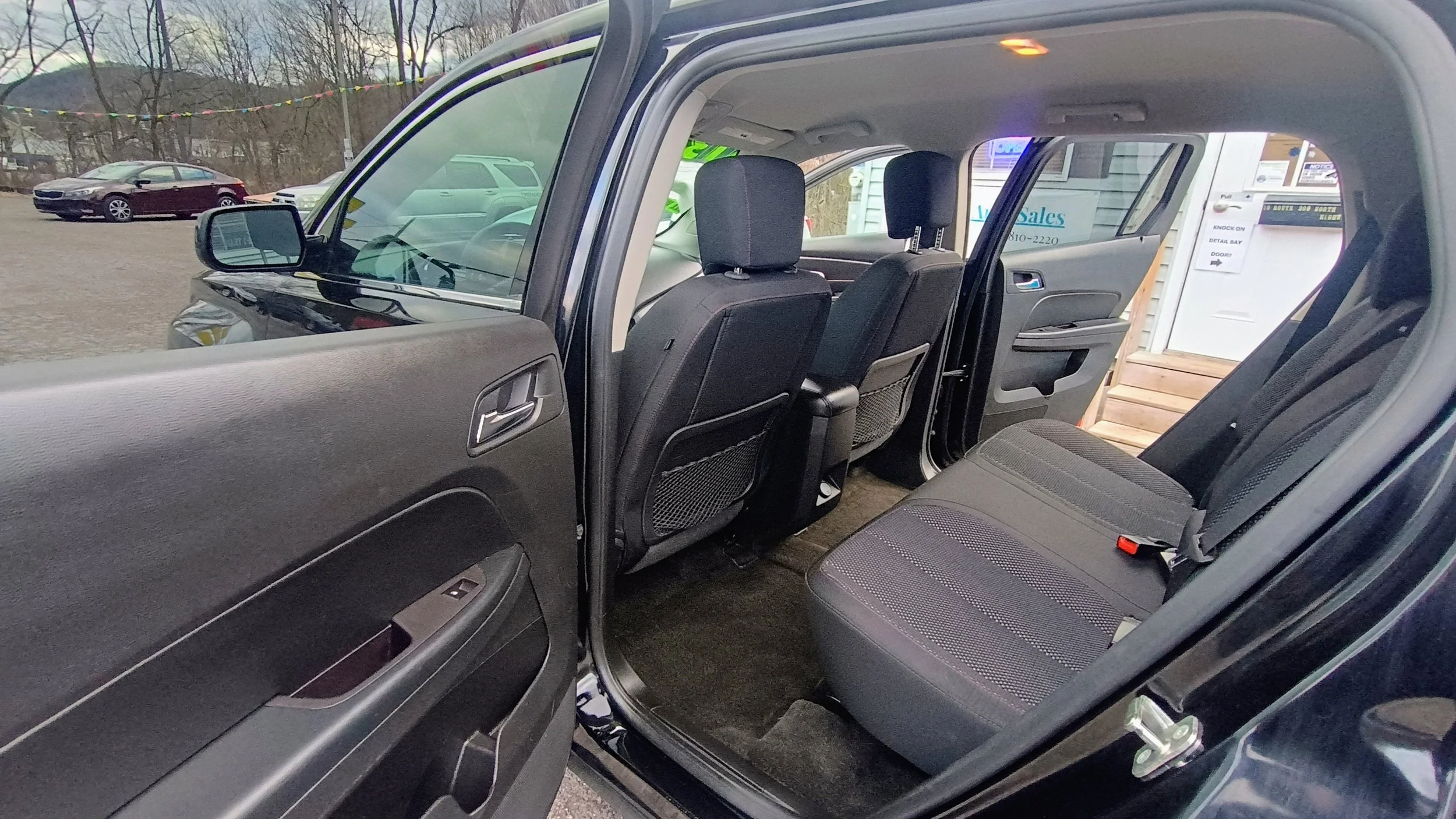 The interior rear passenger area of a black SUV, showing the back seat, seatbelts, and the back of the front seats with mesh storage pockets, parked in a lot near a building with signs and other vehicles in the background.