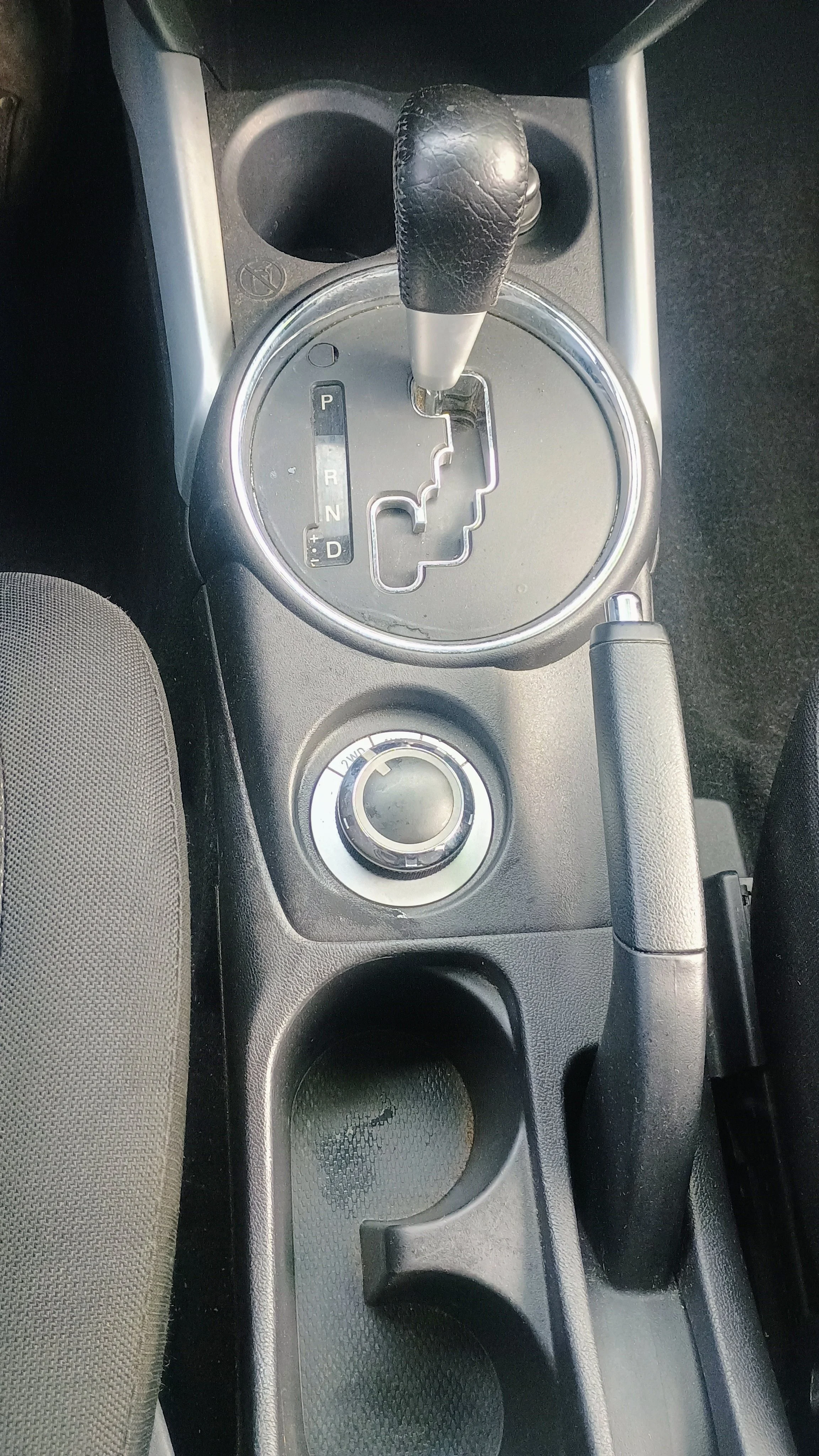 The image shows the center console of a car, featuring a manual gear shift, a rotary dial likely for driving modes, and two cup holders.