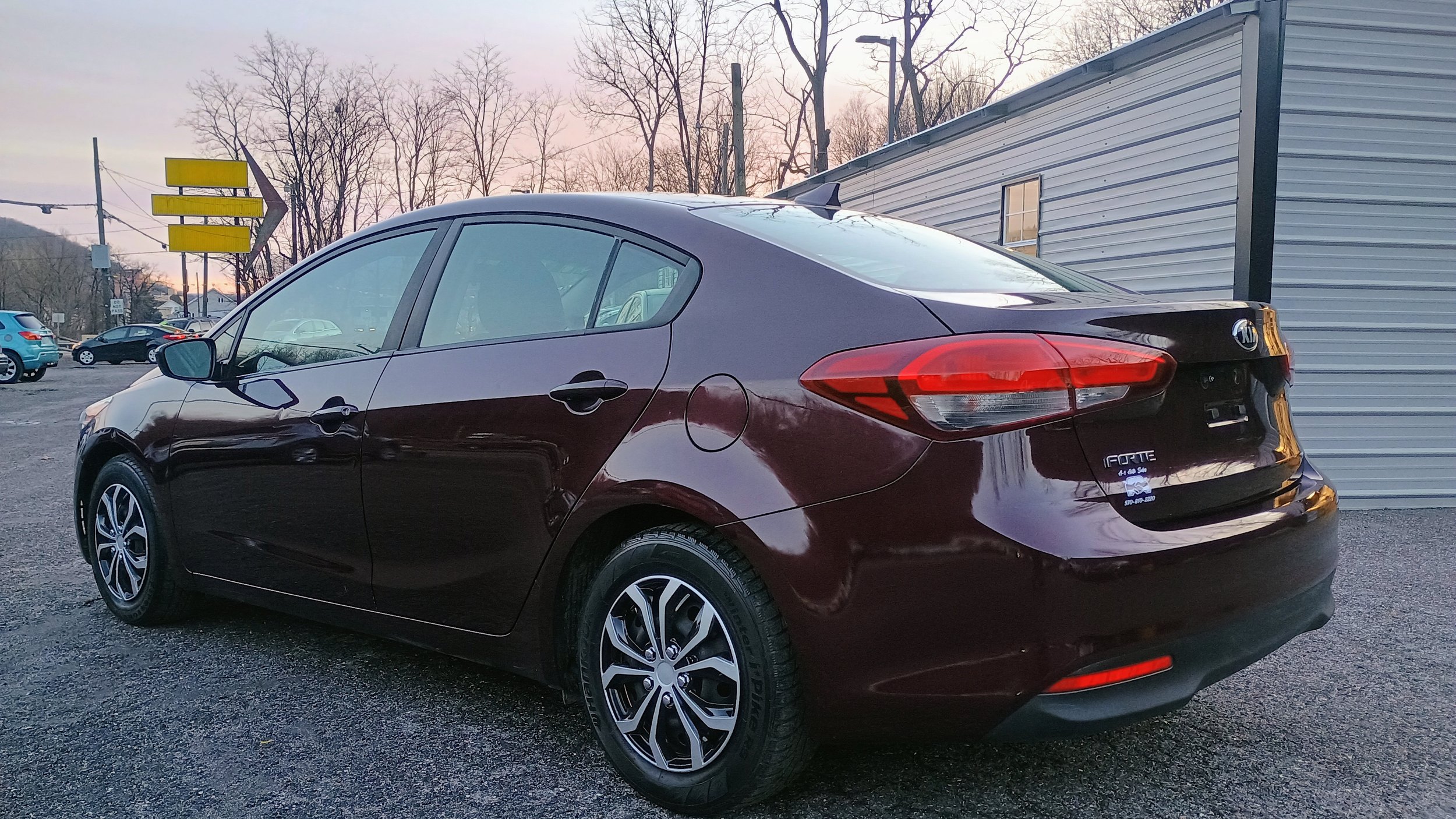 Maroon Kia Forte sedan parked in a gravel lot during sunset with bare trees and other vehicles in the background.