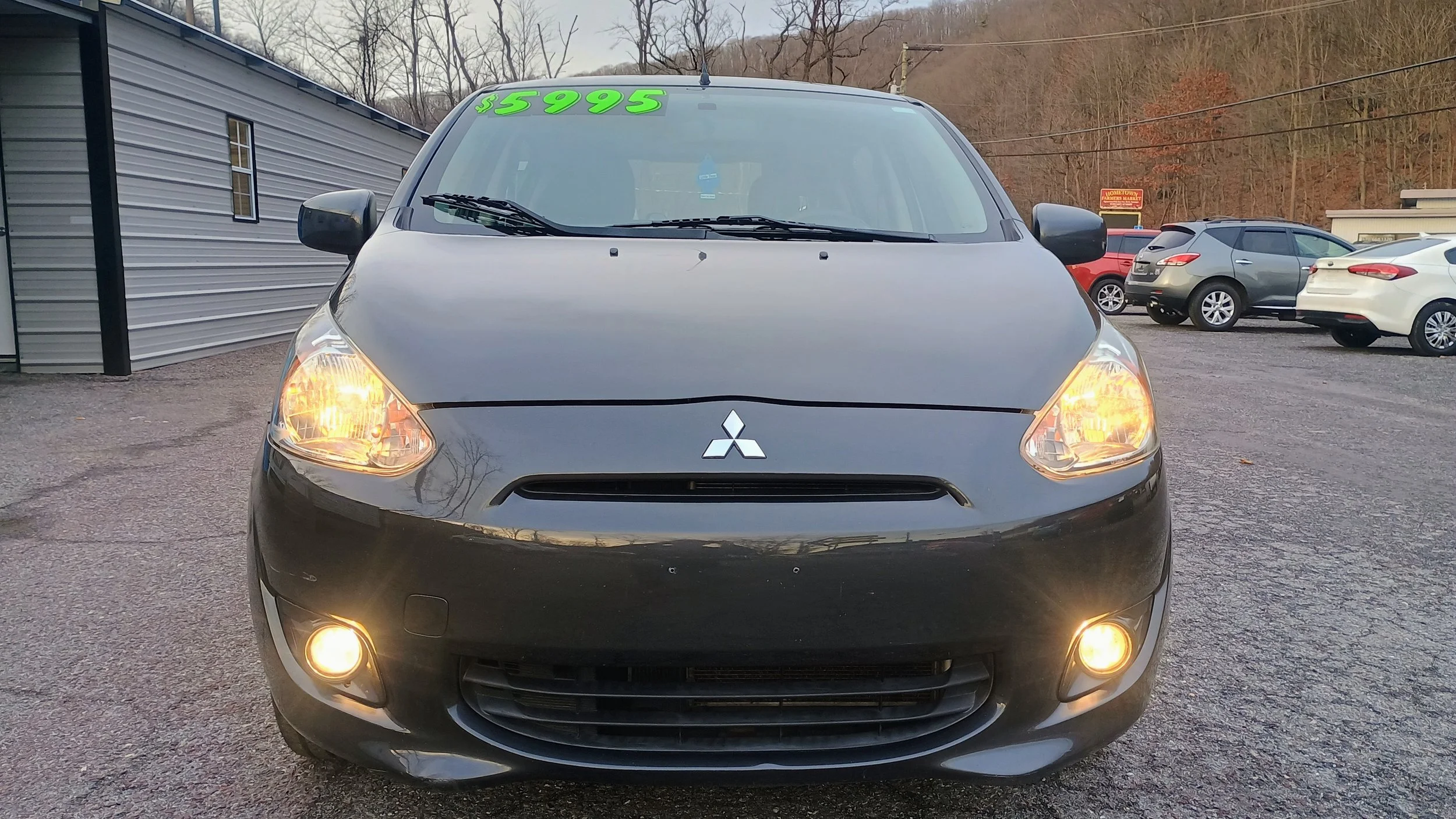 Front view of a black Mitsubishi car with its headlights and fog lights on, displayed for sale at a dealership, with a green price tag of $5995 on the windshield.