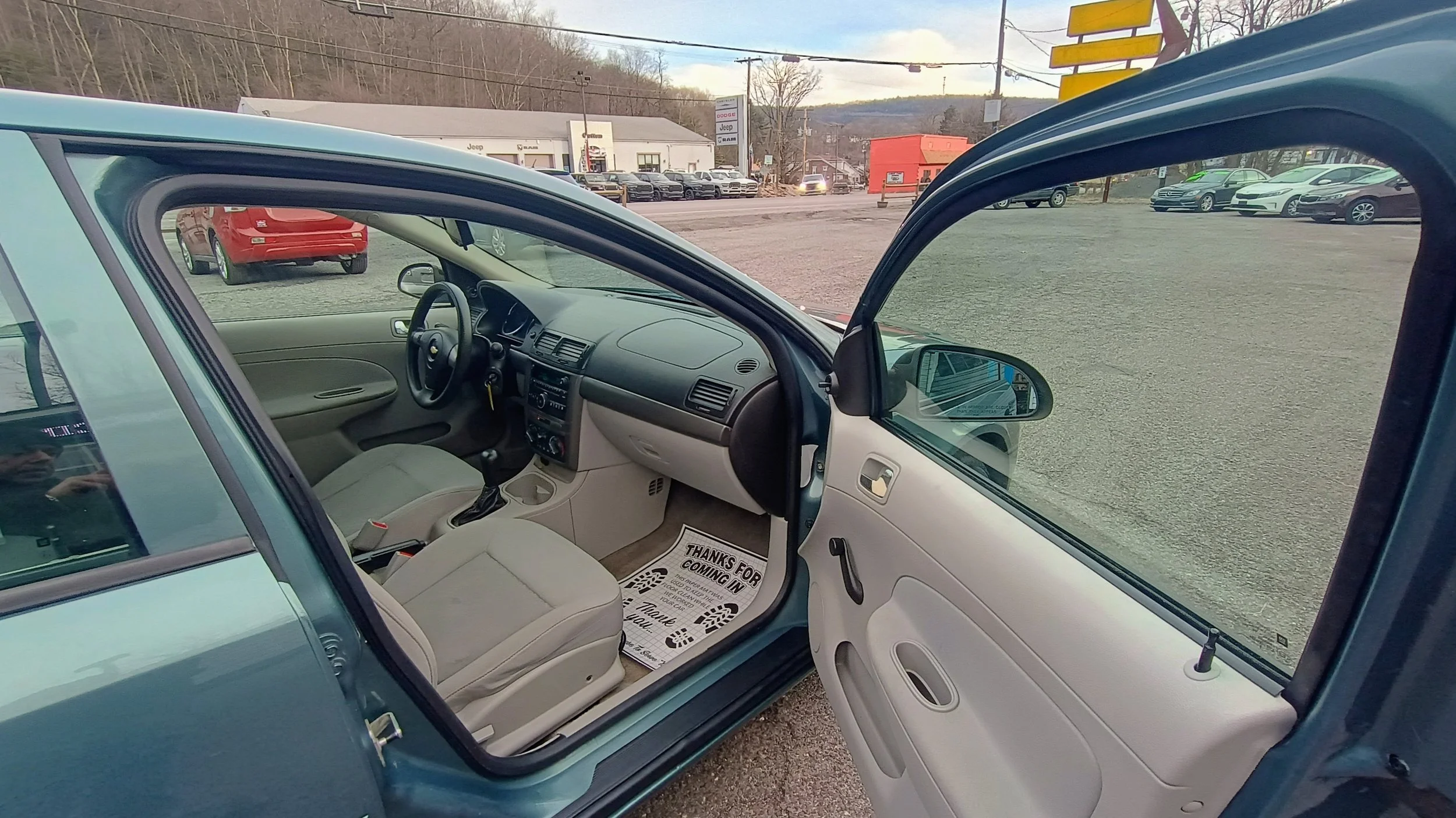 Interior of a small blue car with a view of the front passenger seat, dashboard, and steering wheel, parked at a car dealership lot.