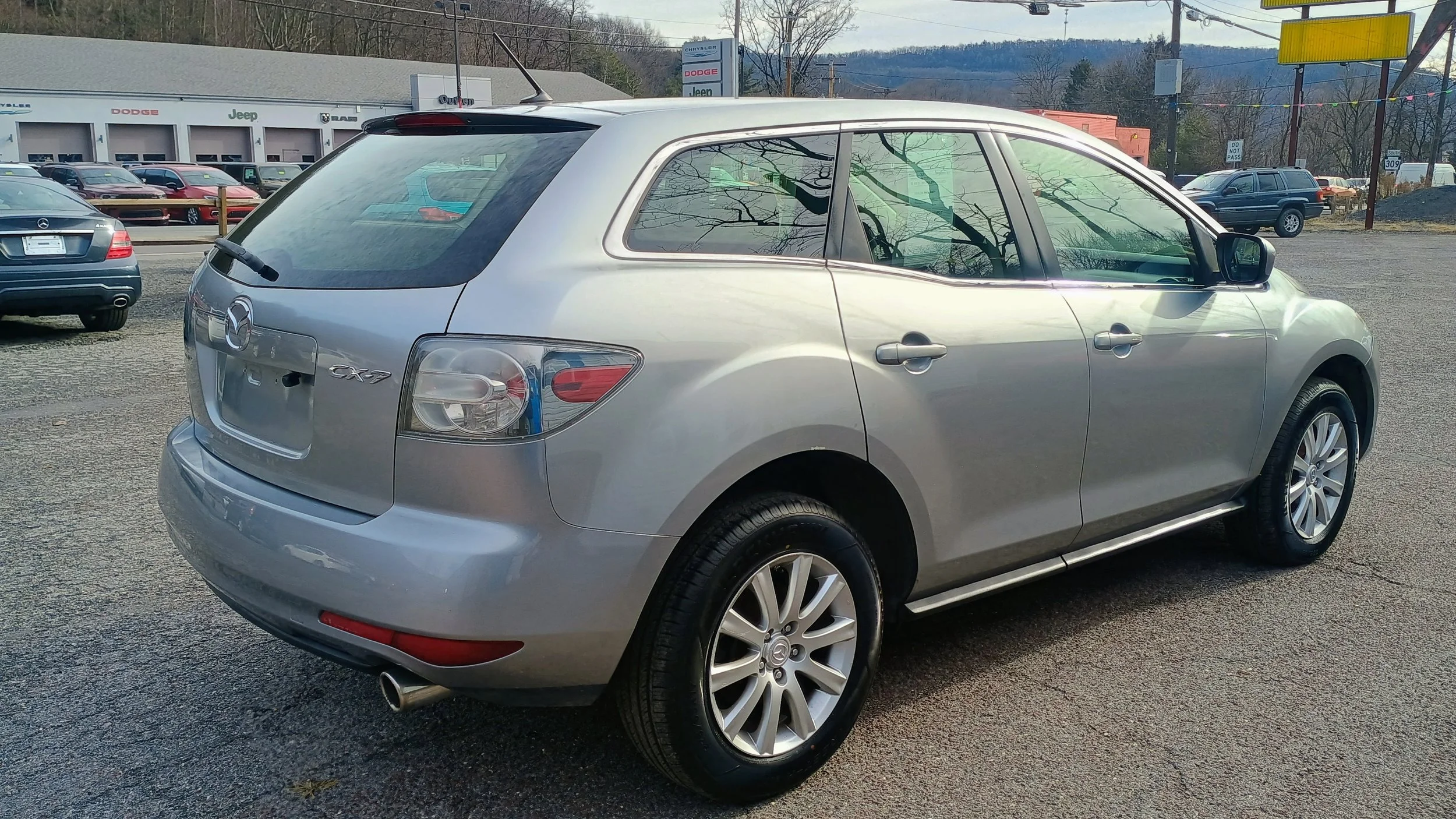 Silver Mazda CX-7 SUV parked in a car dealership lot with other cars and dealership building in the background.