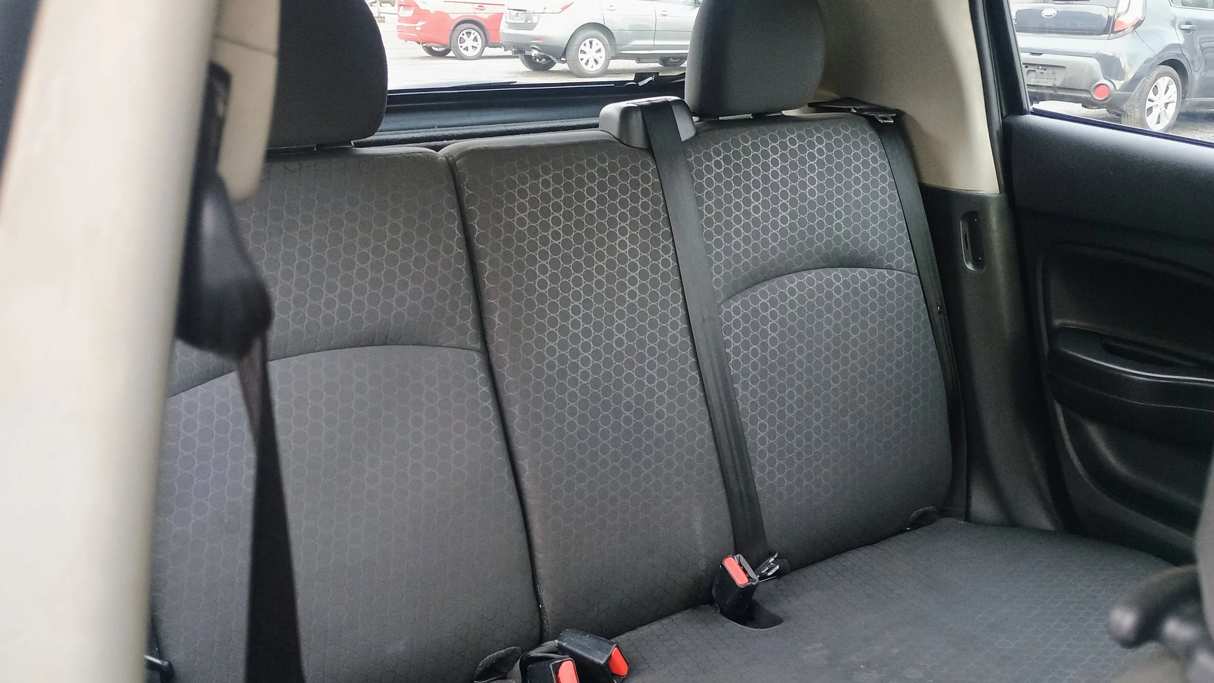 The backseat of a car with three black patterned fabric seats and a seatbelt hanging draped over the middle seat. A window shows parked cars outside.