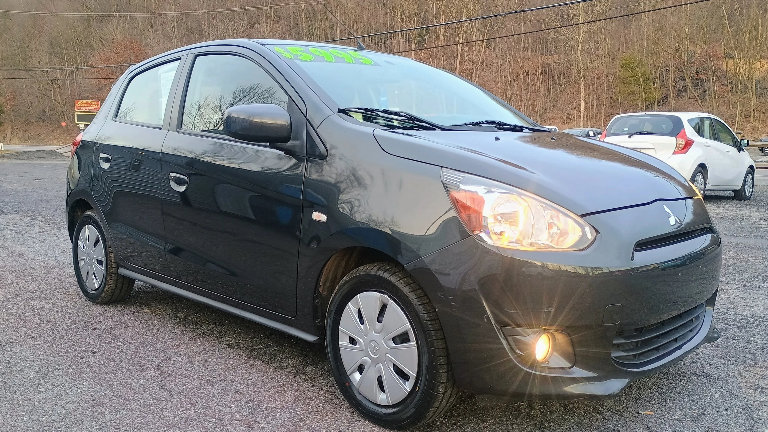 Black Mitsubishi Mirage hatchback with front fog lights on, browsing price $5995, parked on gravel lot with trees in the background.