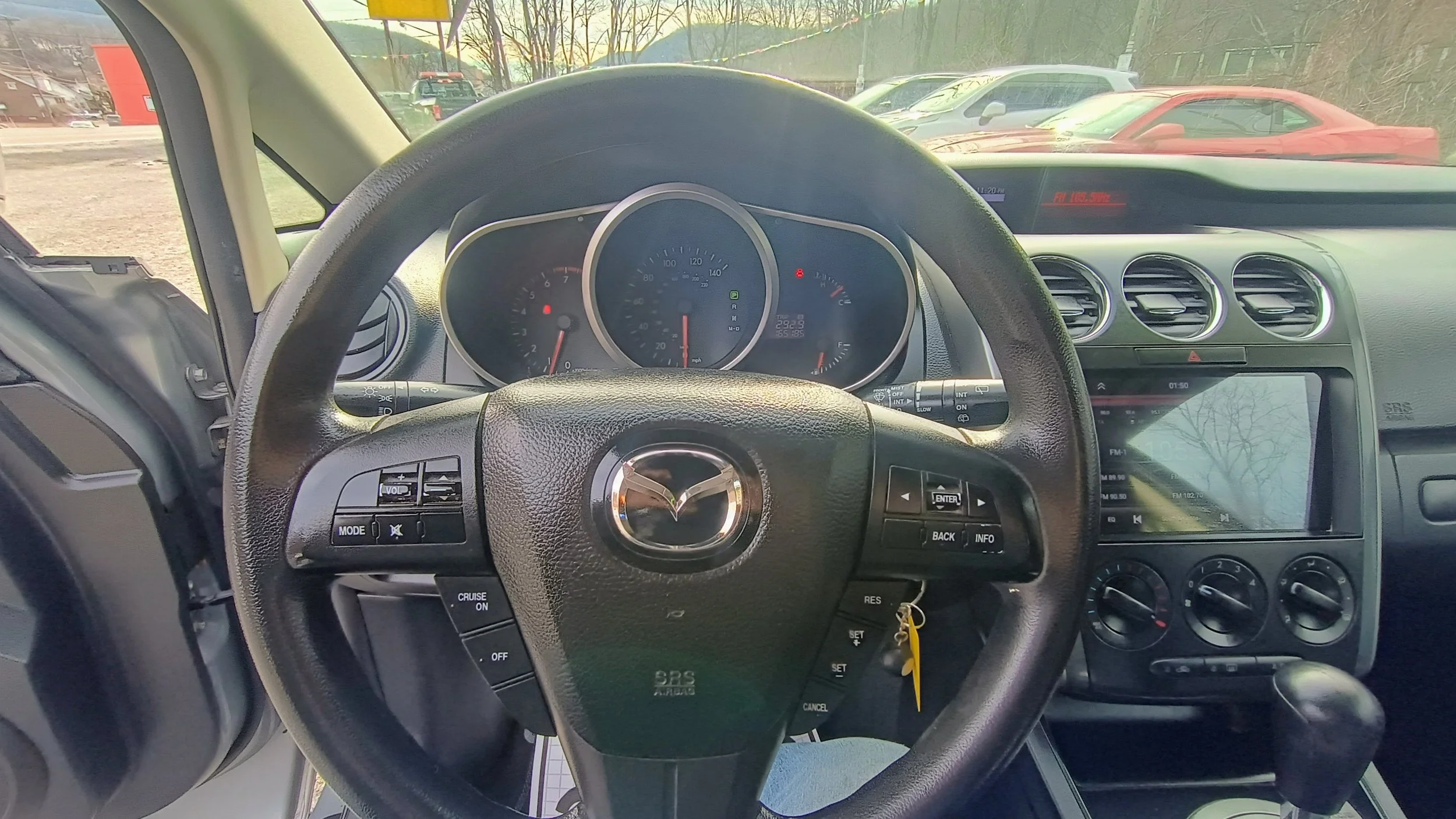 Interior of a car showing the steering wheel, dashboard, and center console. The steering wheel has a Mazda emblem, and the dashboard displays the speedometer, tachometer, and fuel gauge. The center console includes air vents, a touchscreen display, and climate control knobs. Outside the windshield, a parking lot with several cars and trees is visible.