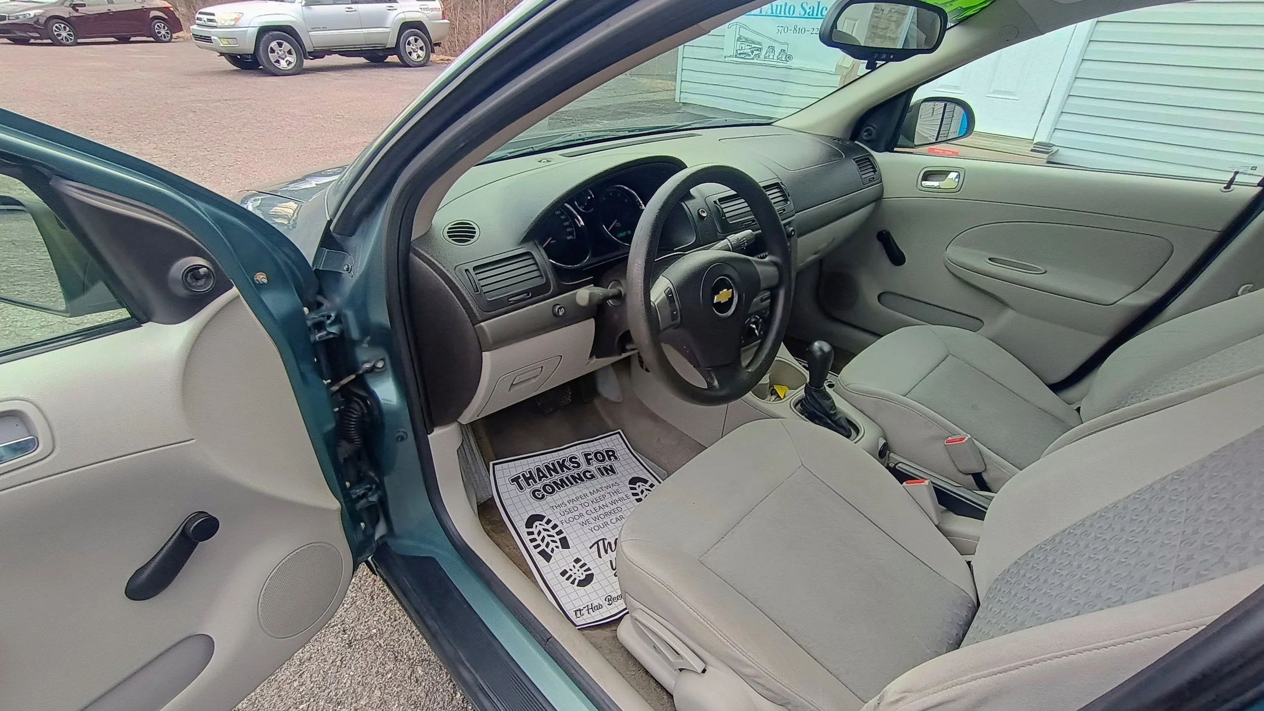 The interior of a Chevrolet vehicle showing the driver's seat, steering wheel, dashboard, and front passenger seat, with a view of parked cars and a building outside.