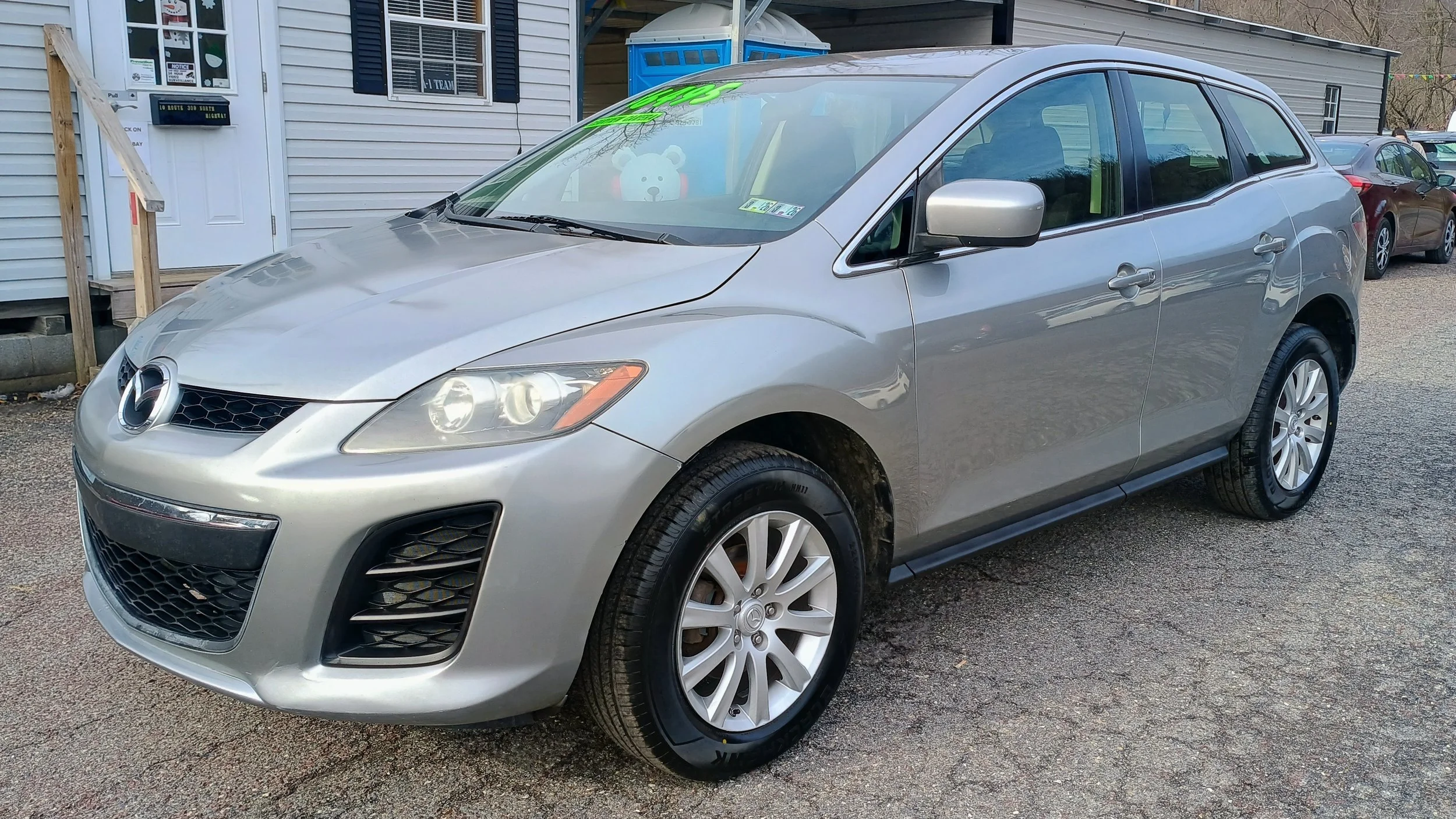 Silver Mazda sedan parked on an asphalt lot in front of a house with white siding. The car has a stuffed animal toy on the dashboard and a green price sticker on the windshield.