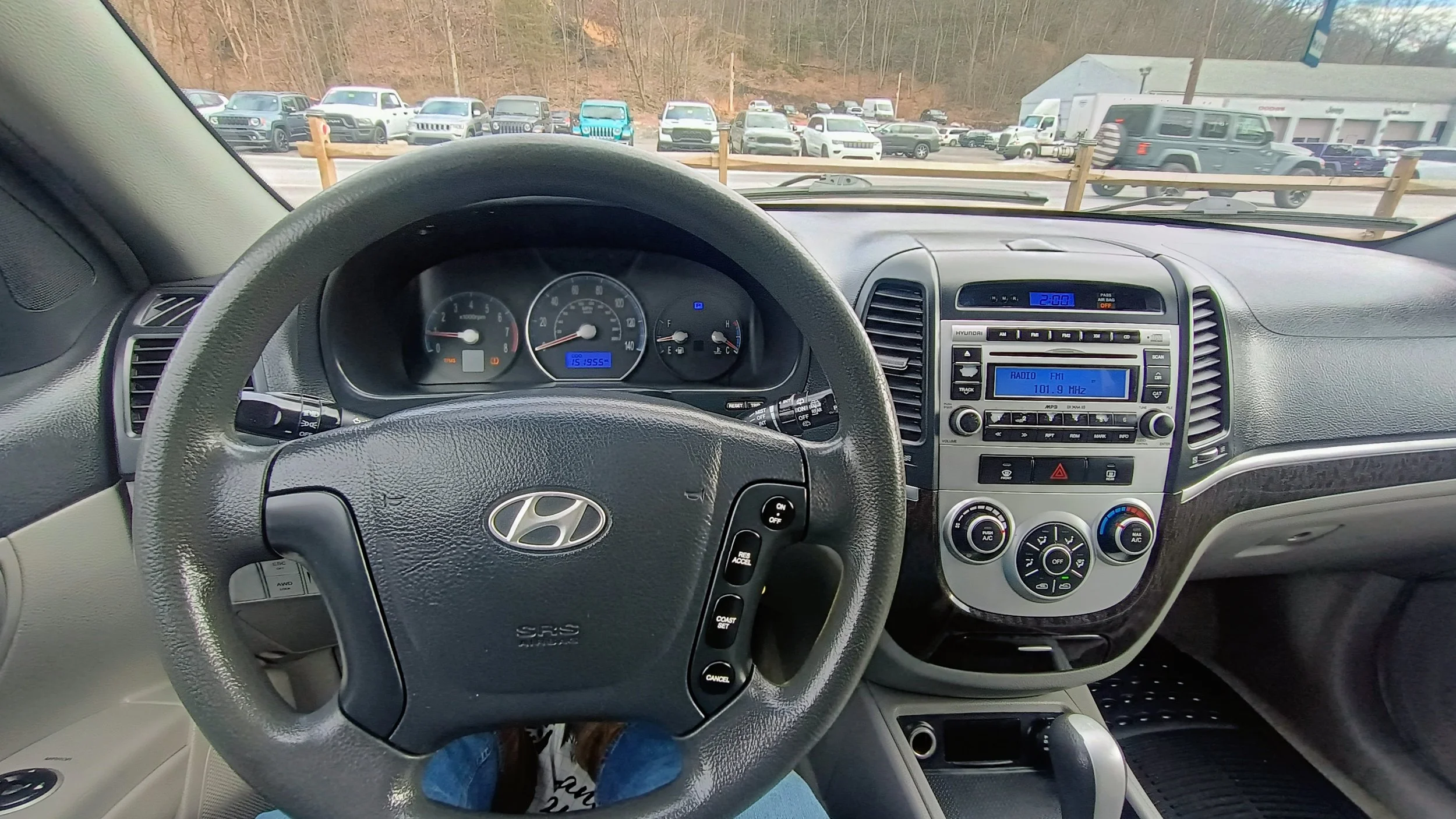 View from inside a Hyundai car showing the dashboard, steering wheel, and parking lot with numerous cars and an overcast sky in the background.