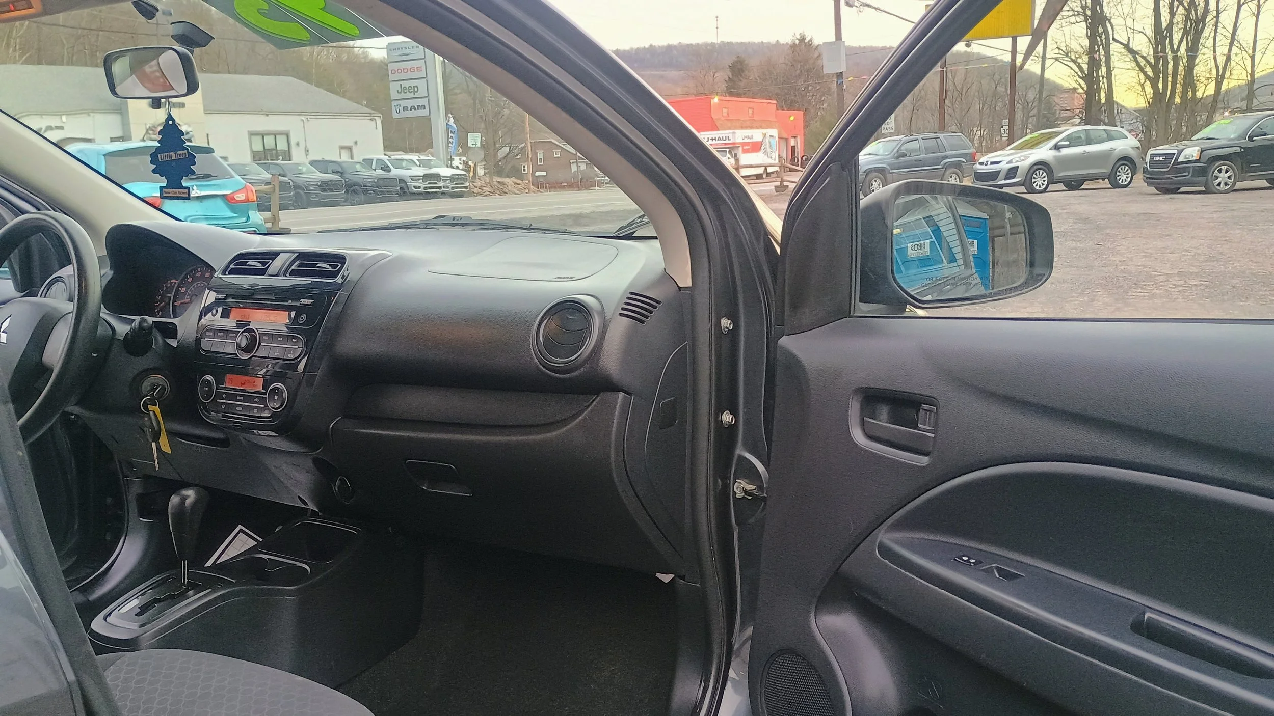 Inside view of a vehicle showing the dashboard, steering wheel, gear shift, and the passenger side door with window controls, with a parking lot and cars visible outside.