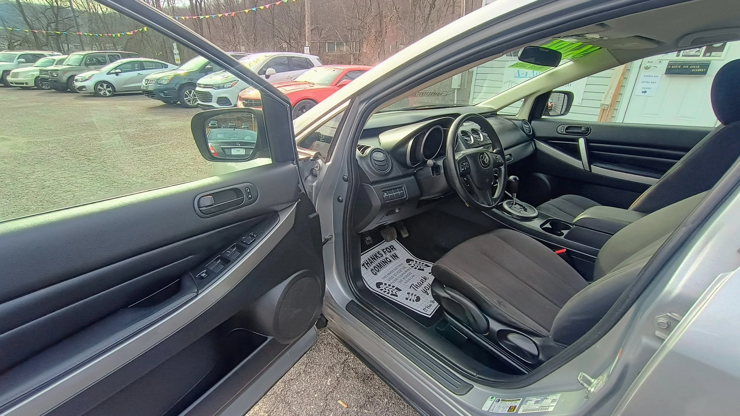 Interior of a right-hand drive car parked with the driver's side open in a car dealership lot, with a multi-colored pennant banner overhead and other cars visible in the background.