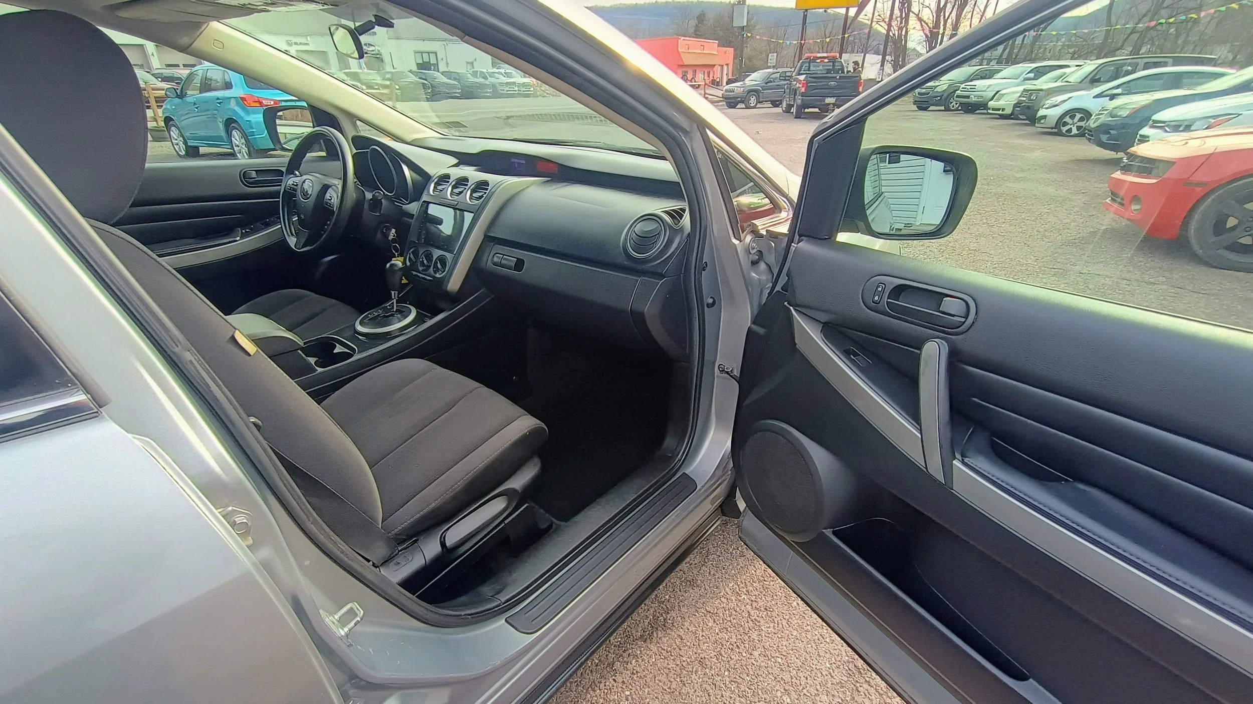 The interior of a silver car with the driver's side door open, showing the driver's seat, dashboard, and steering wheel, with a view of a parking lot filled with various parked cars in the background.