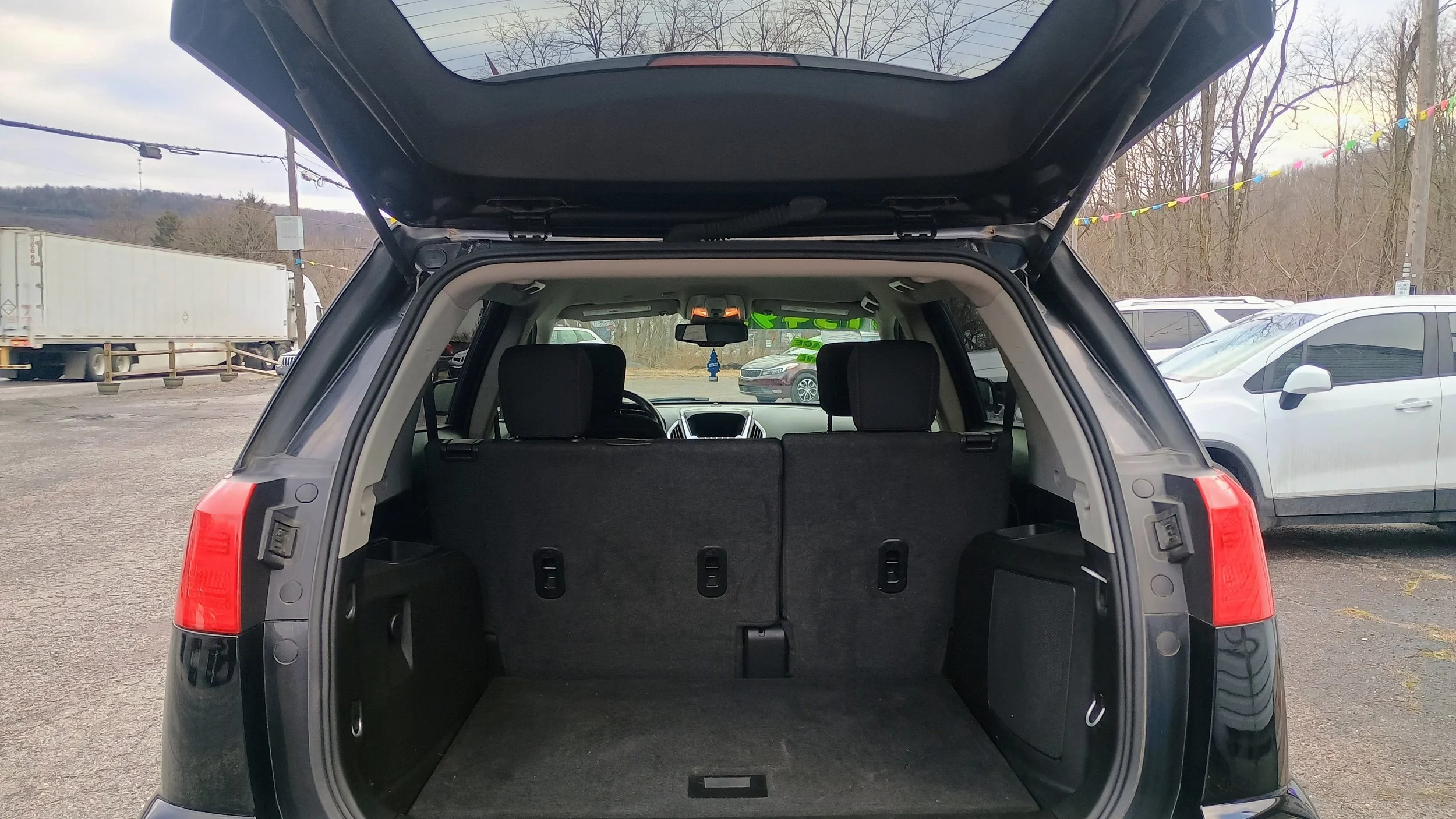 Rear view of an SUV with its hatch open, showing the cargo area with folded rear seats, in a parking lot with other vehicles and trees in the background.
