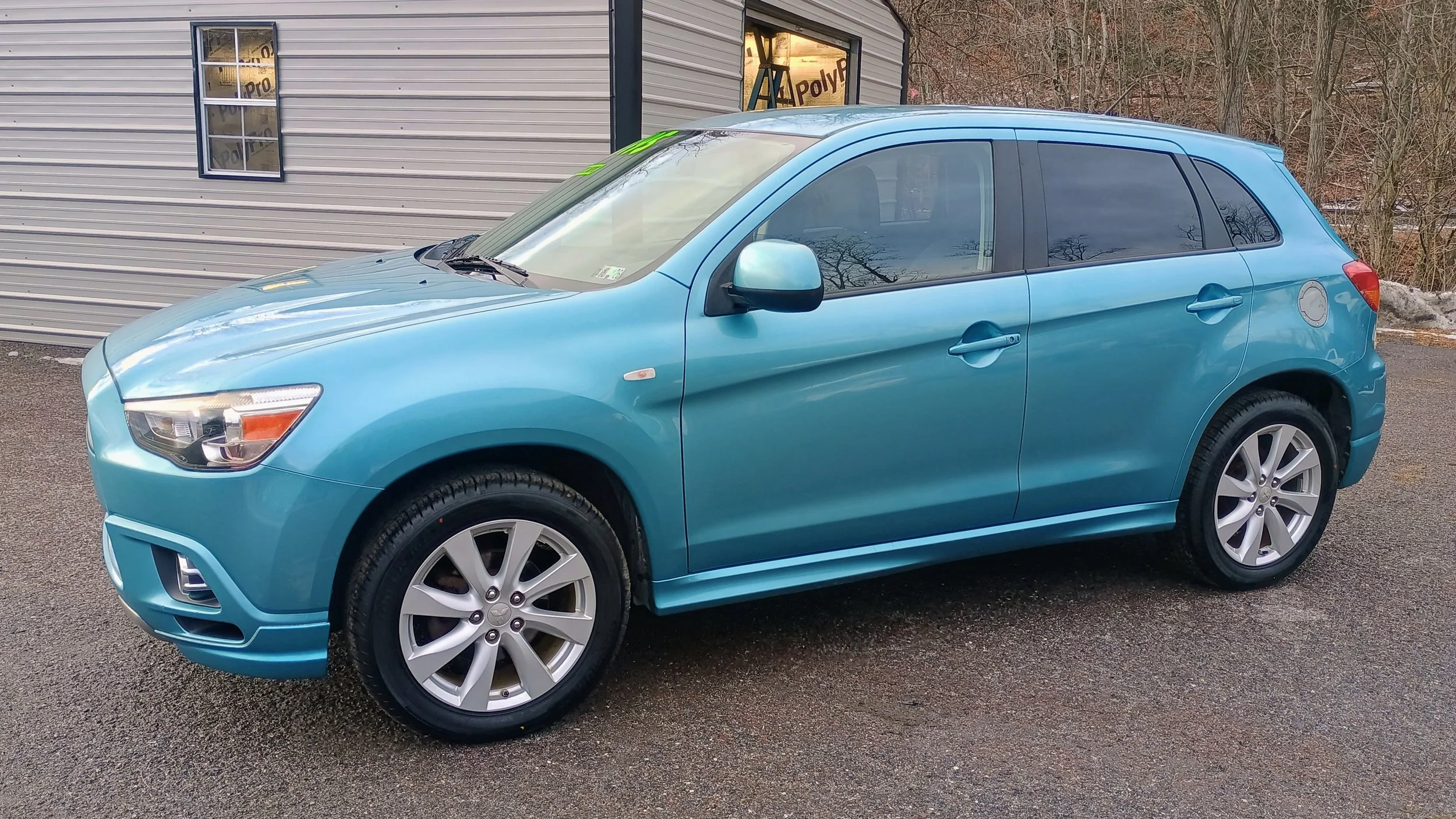 A blue compact hatchback car parked on asphalt near a building with windows and a wooded area in the background.