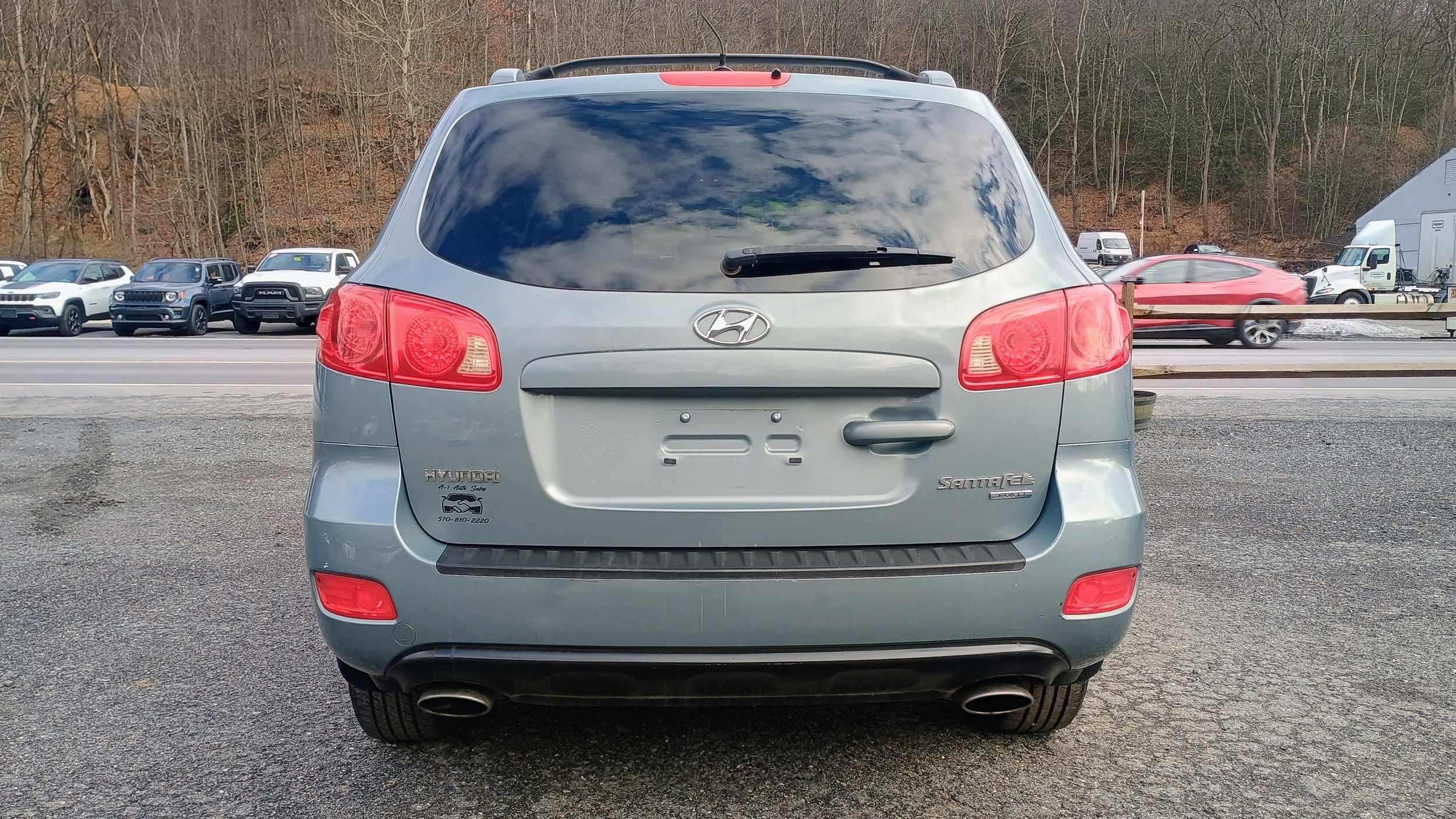 Gray Hyundai Santa Fe SUV parked in a lot, with a line of vehicles and a wooded hill in the background.