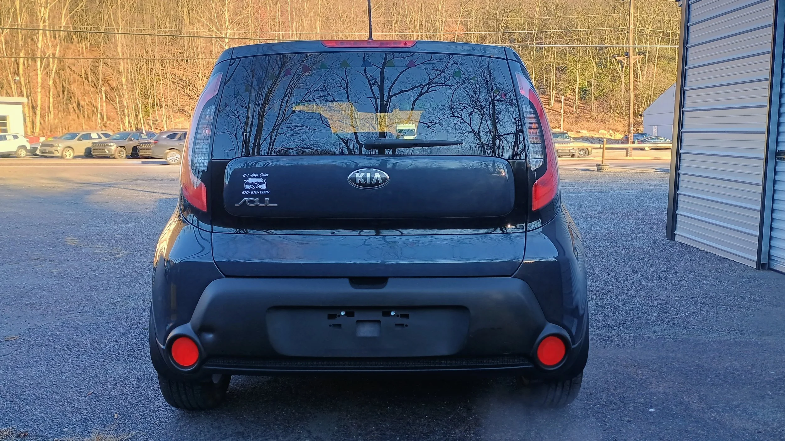 Rear view of a black Kia Soul parked on pavement with a license plate holder, taillights, and reflective orange reflectors, with a parking lot and trees in the background.