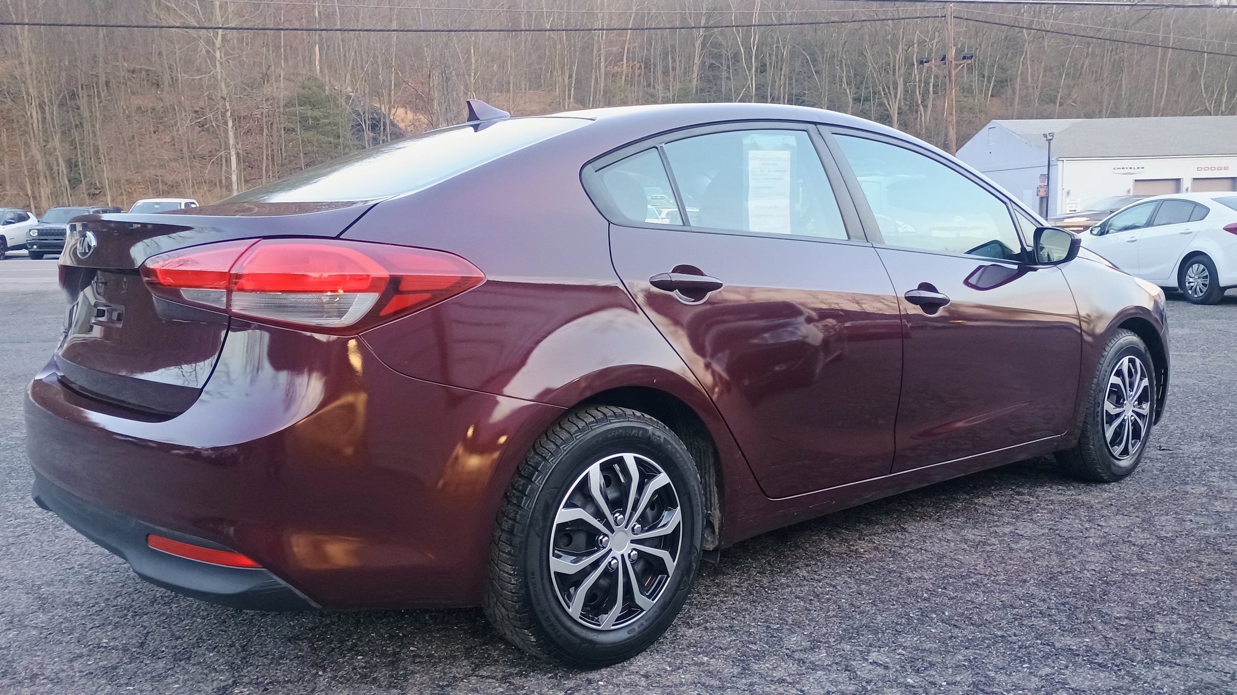 Rear and side view of a dark purple sedan car parked outdoors on a gravel surface, with other vehicles and a wooded hill in the background.