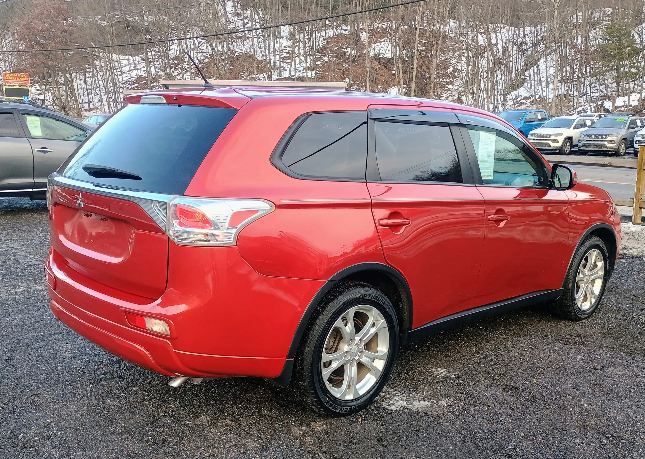 Red station wagon parked on a gravel lot with other cars in the background, snow on the ground and trees without leaves.