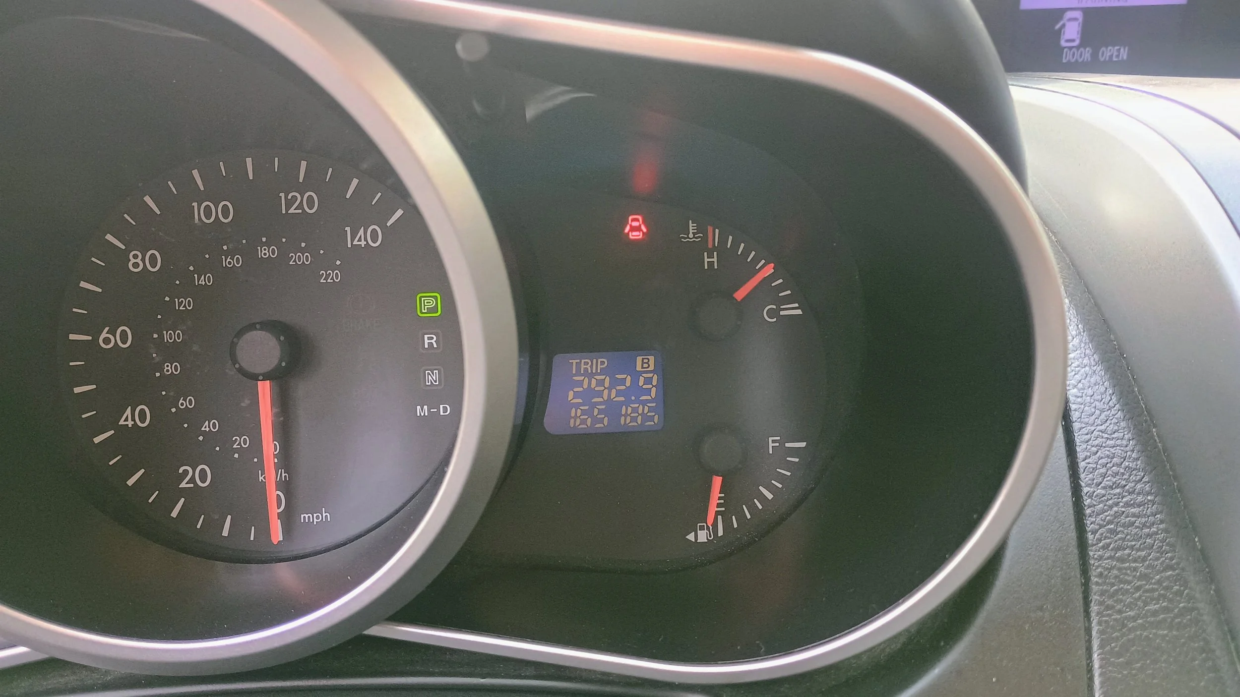 Car dashboard showing the speedometer at 0 mph, fuel gauge nearly empty, trip meter at 292.9 miles, and a warning light for the seat belt indicator is on. The background shows the outside is bright.