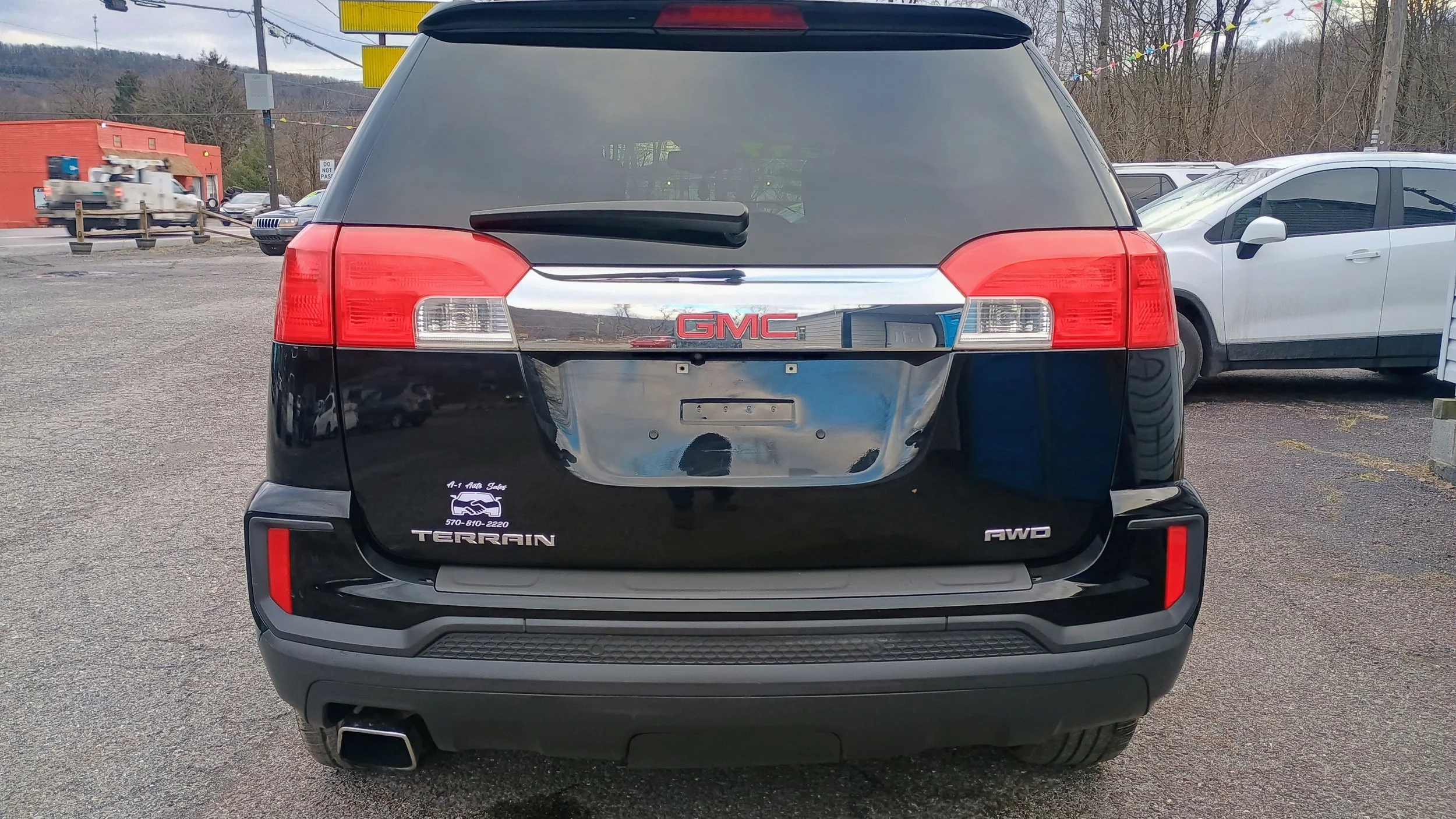 Rear view of a black GMC Terrain SUV with a missing license plate, parked in a lot with other vehicles, including a white car to the right. The vehicle has decals that read 'T-A Auto Sales' and 'AWD'.