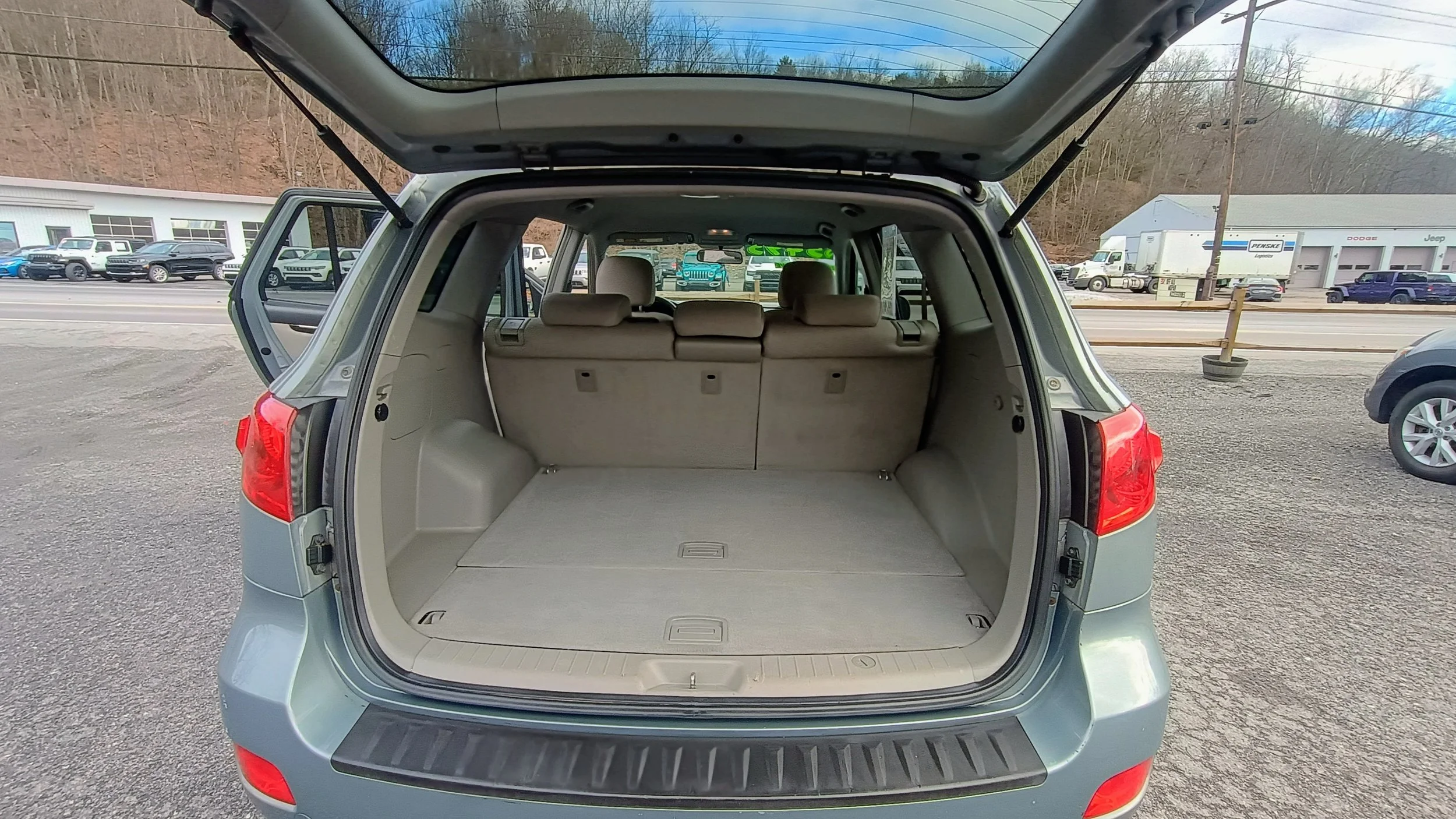 Open rear hatch of a silver SUV showing an empty cargo area with beige interior and three rear seats. Outside, a car dealership lot with various parked cars and a dealership building in the background.