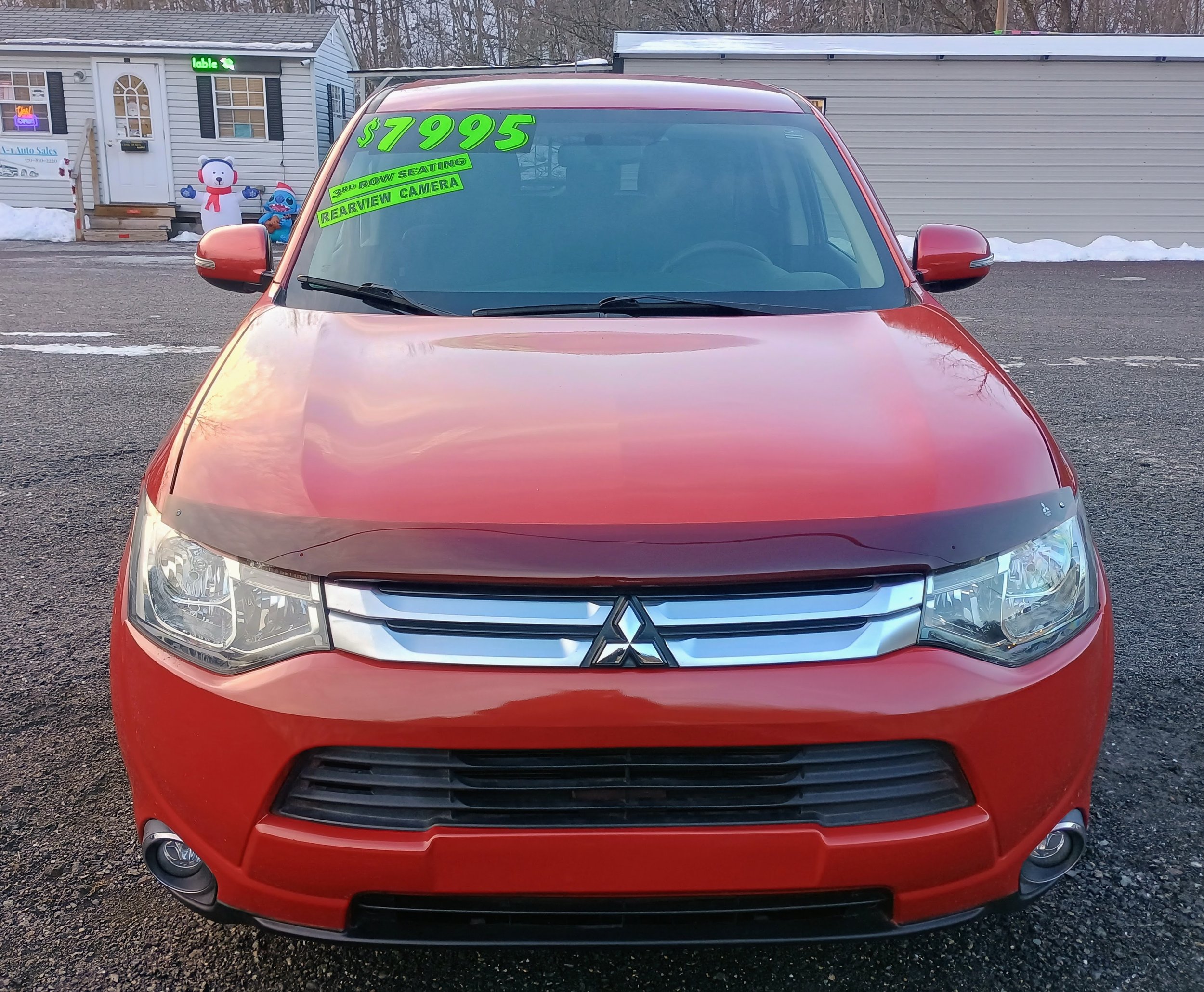 Red Mitsubishi SUV parked in a lot with sale signs on the windshield indicating the price of $7995, third-row seating, and a rearview camera.
