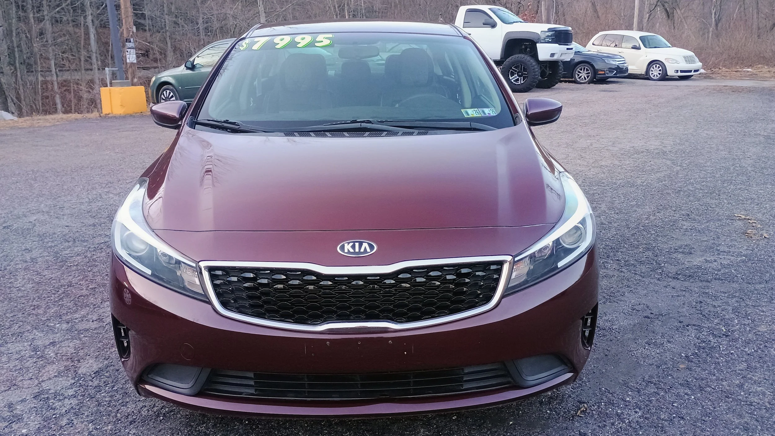 Maroon Kia sedan parked in a gravel lot with other cars and trucks in the background, hill with trees behind.