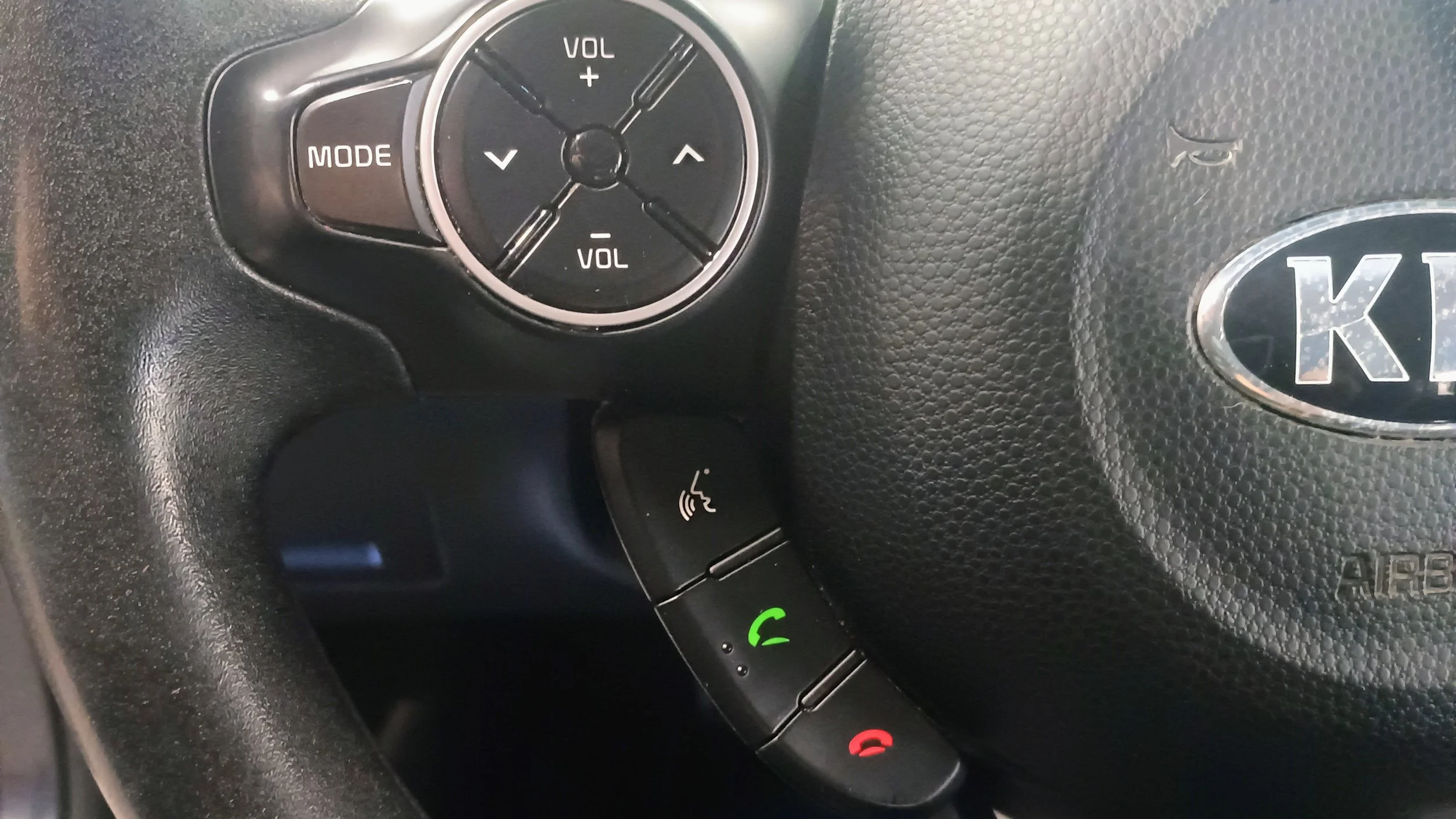 Close-up of a steering wheel in a Kia vehicle with integrated controls, including a circular button with volume and mode controls, and a series of buttons for answering and ending calls, with a black textured surface and the Kia logo.