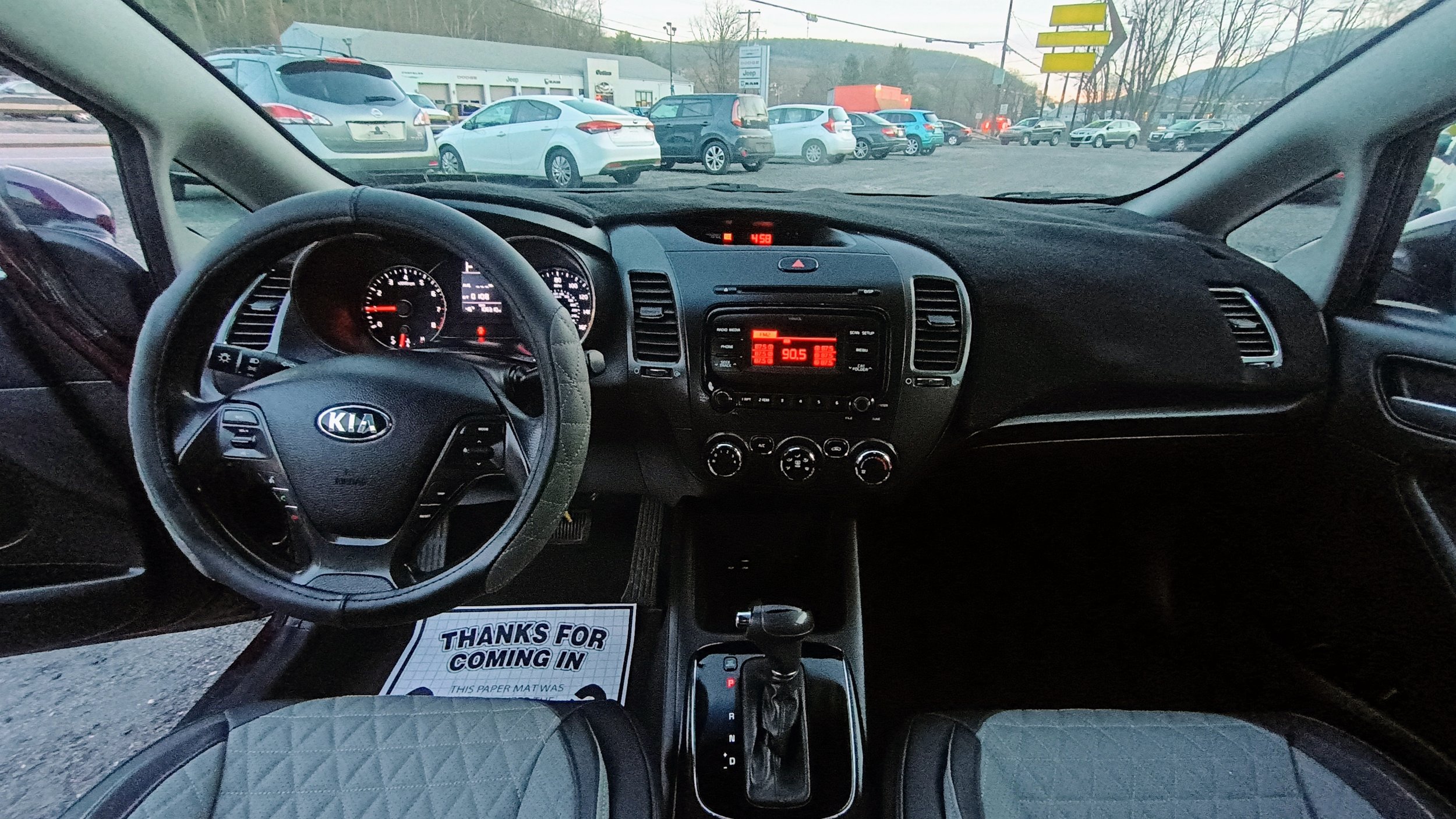Interior view of a Kia car dashboard and steering wheel, with the parking lot and other cars visible through the windshield.