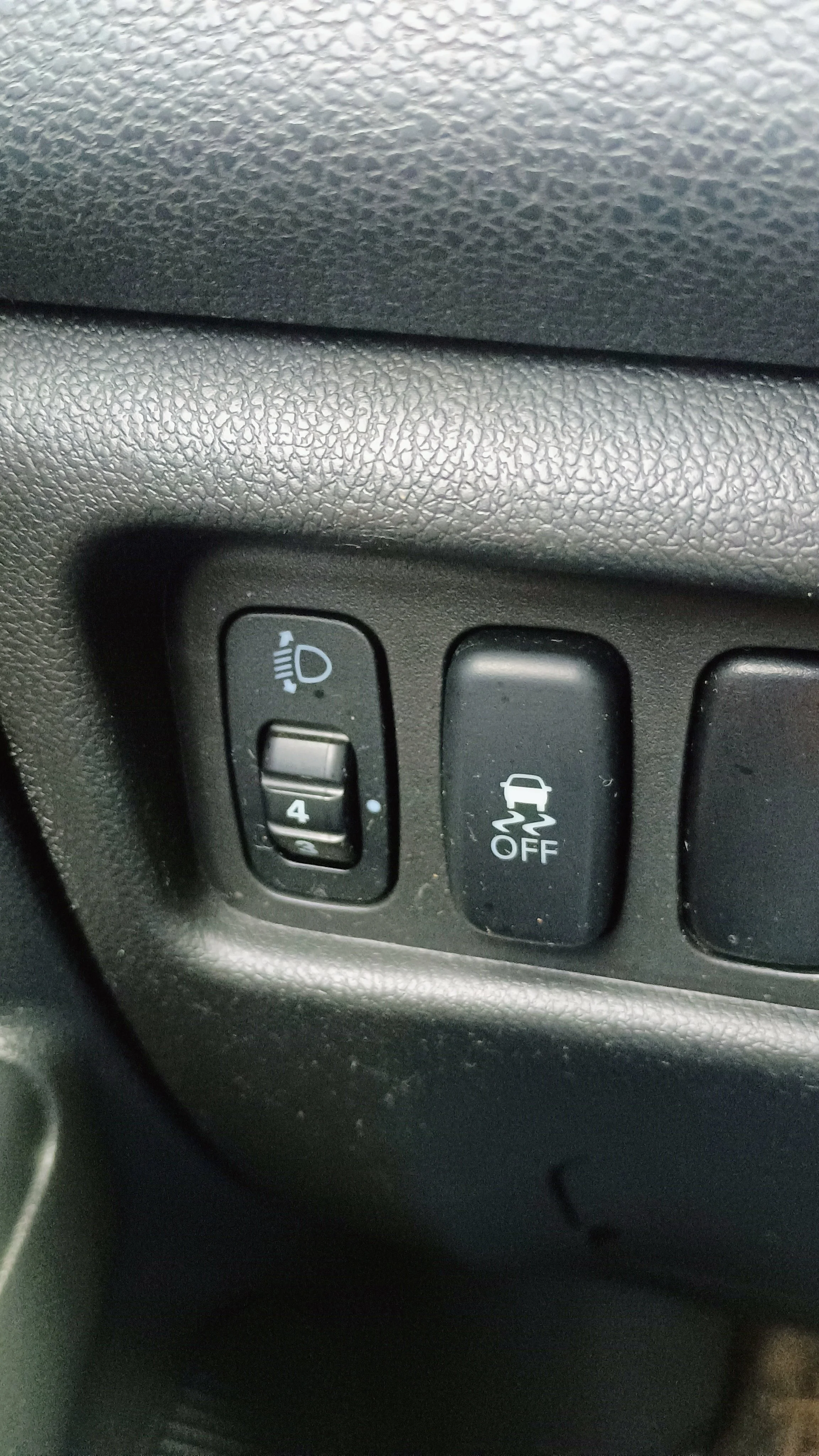 Close-up of a car dashboard showing headlight adjustment switch, vehicle stability control off button, and dashboard textured surface.