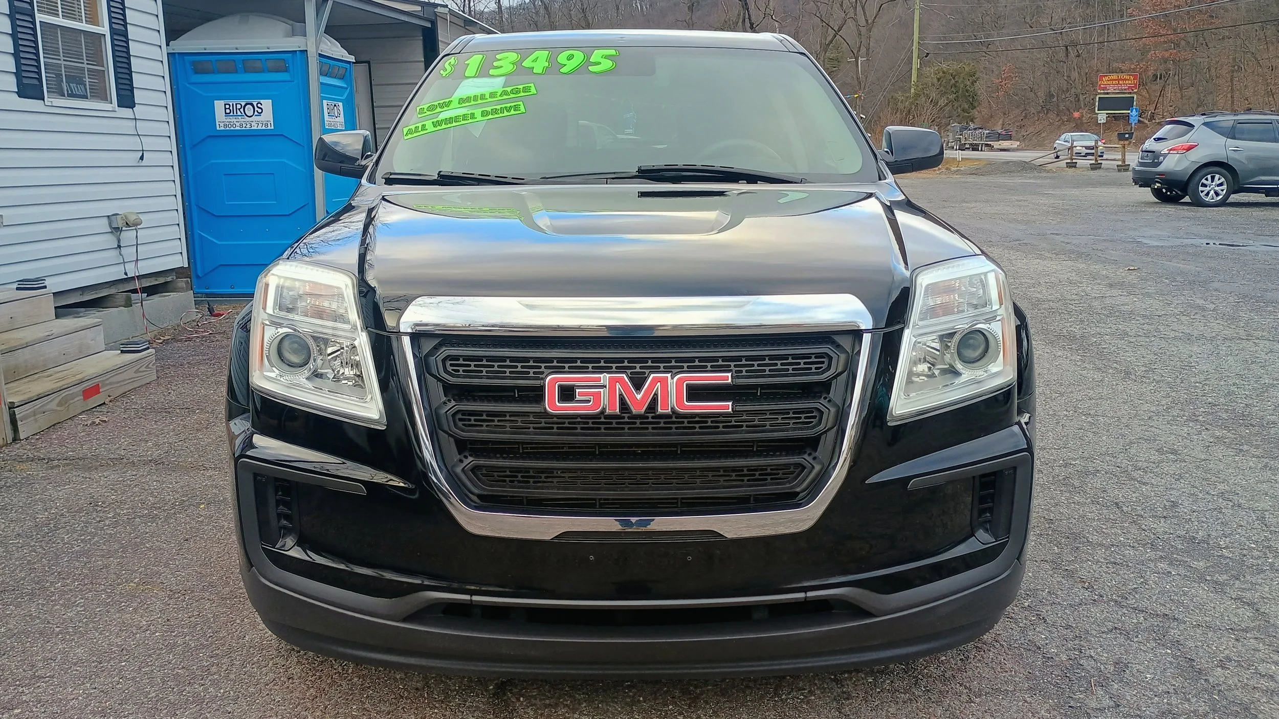 Front view of a black GMC SUV for sale, with green price and details on the windshield, parked on a gravel lot near a building and other vehicles in the background.