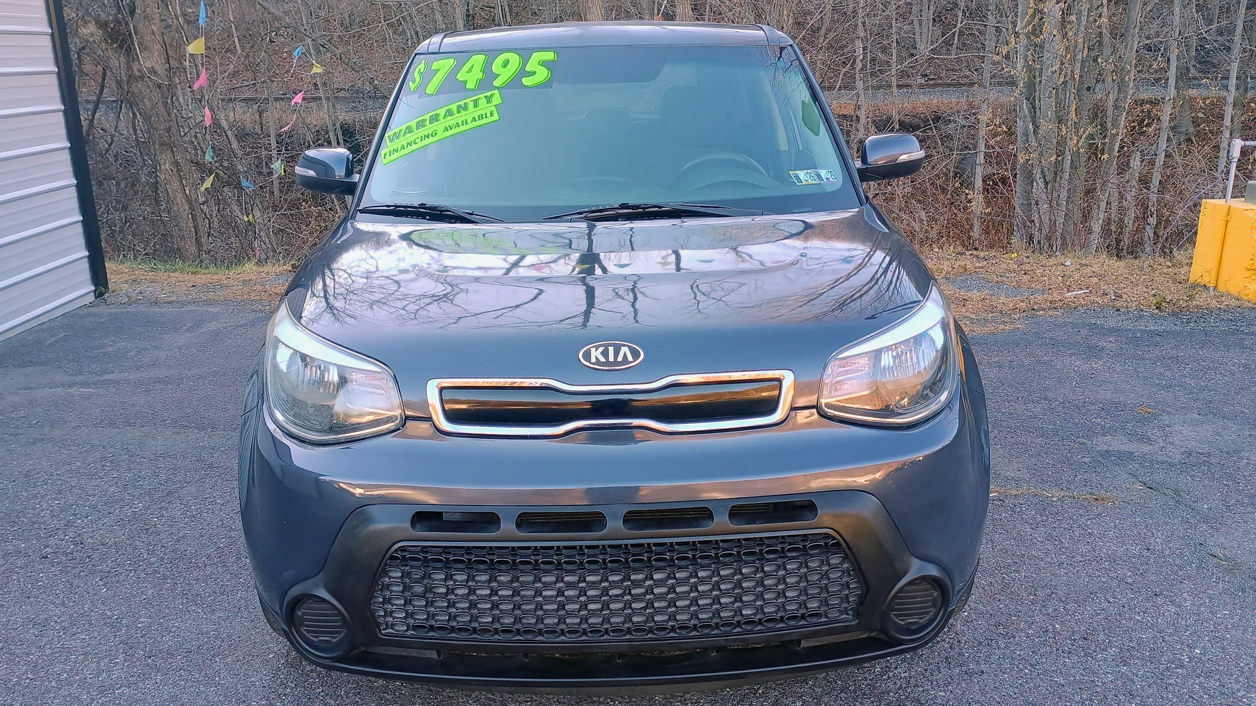 Black Kia car for sale at a dealership, with a price of $7495 displayed on the windshield, and a green sticker indicating warranty and financing options.