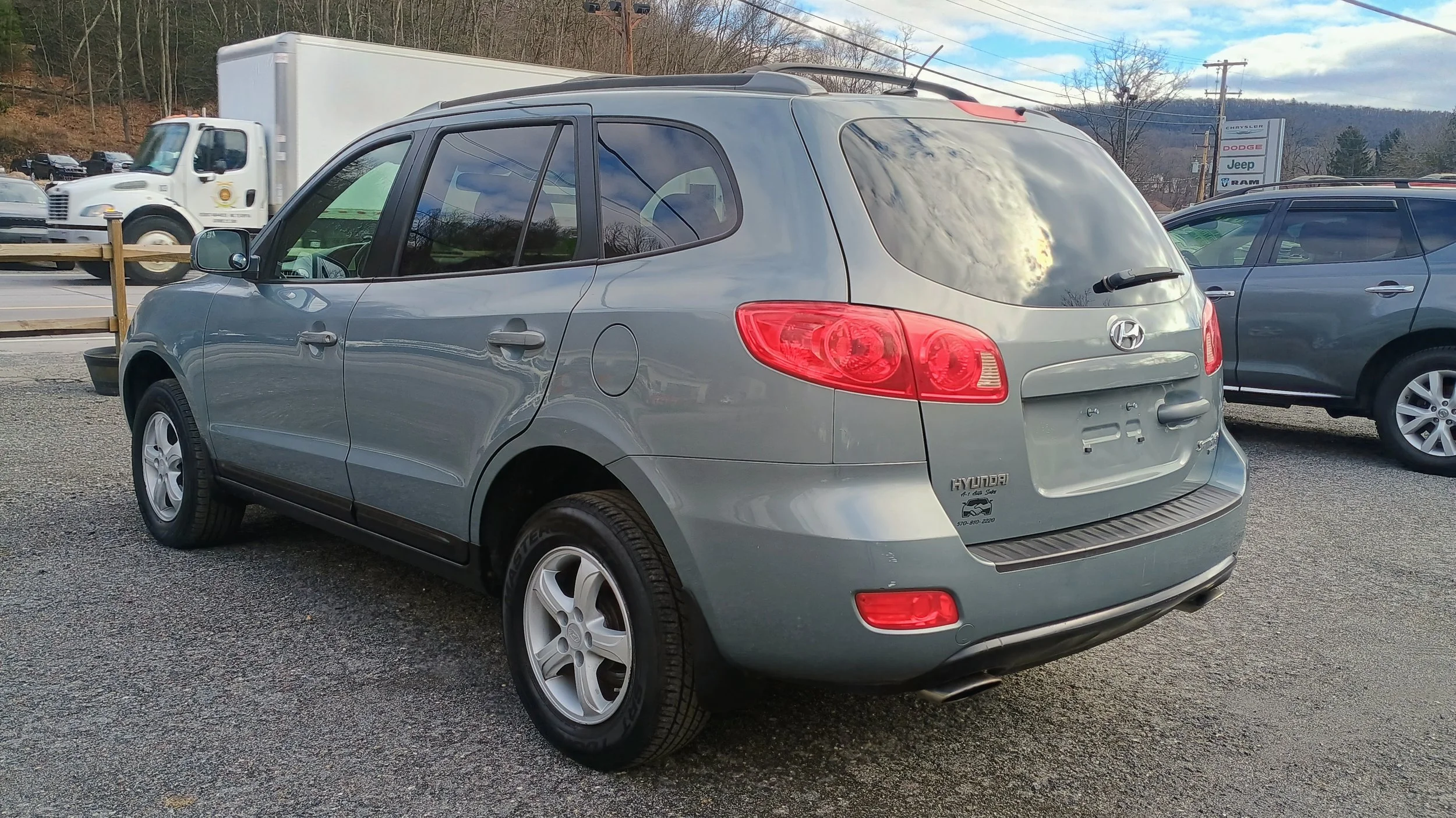 Gray Hyundai Tucson parked in a lot with other vehicles and a dealership sign in the background, cloudy sky reflected in the rear window.
