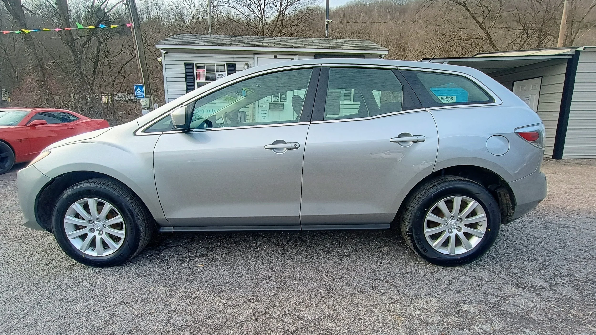 Side view of a silver compact hatchback car parked on asphalt, with buildings and other vehicles in the background.