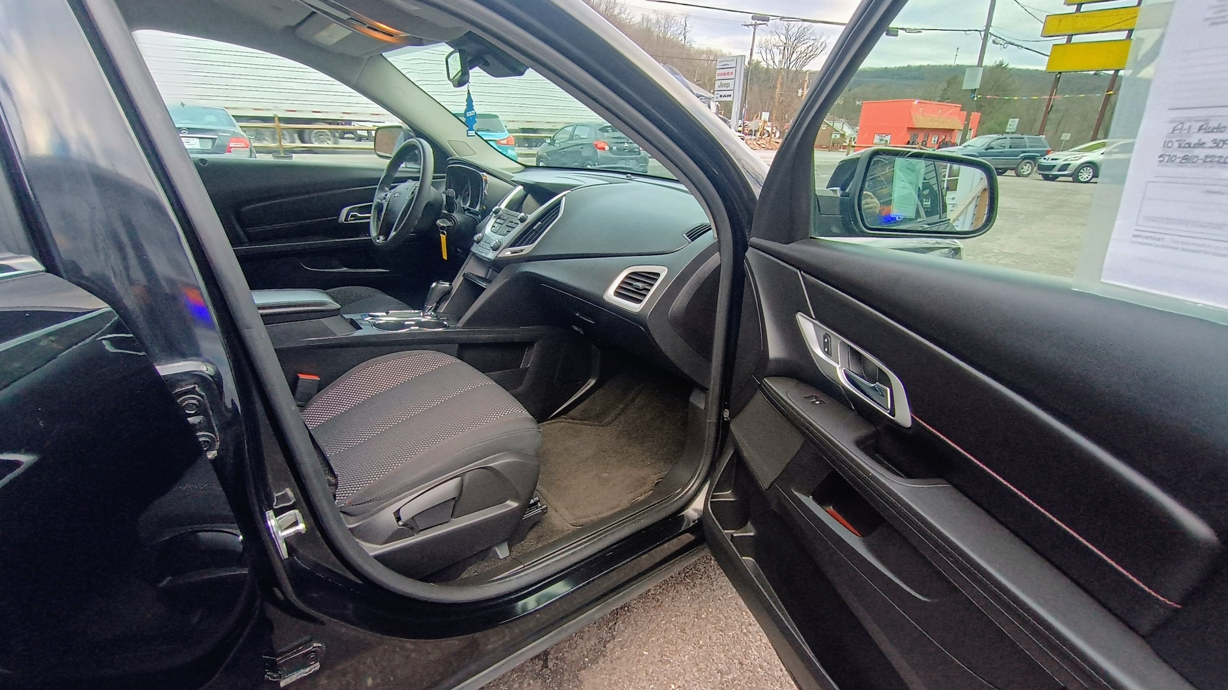 Interior of a black SUV showing the front passenger seat, dashboard, steering wheel, and open front passenger door. The driver’s seat and part of the rear seats are also visible, with vehicles and a dealership lot outside the window.