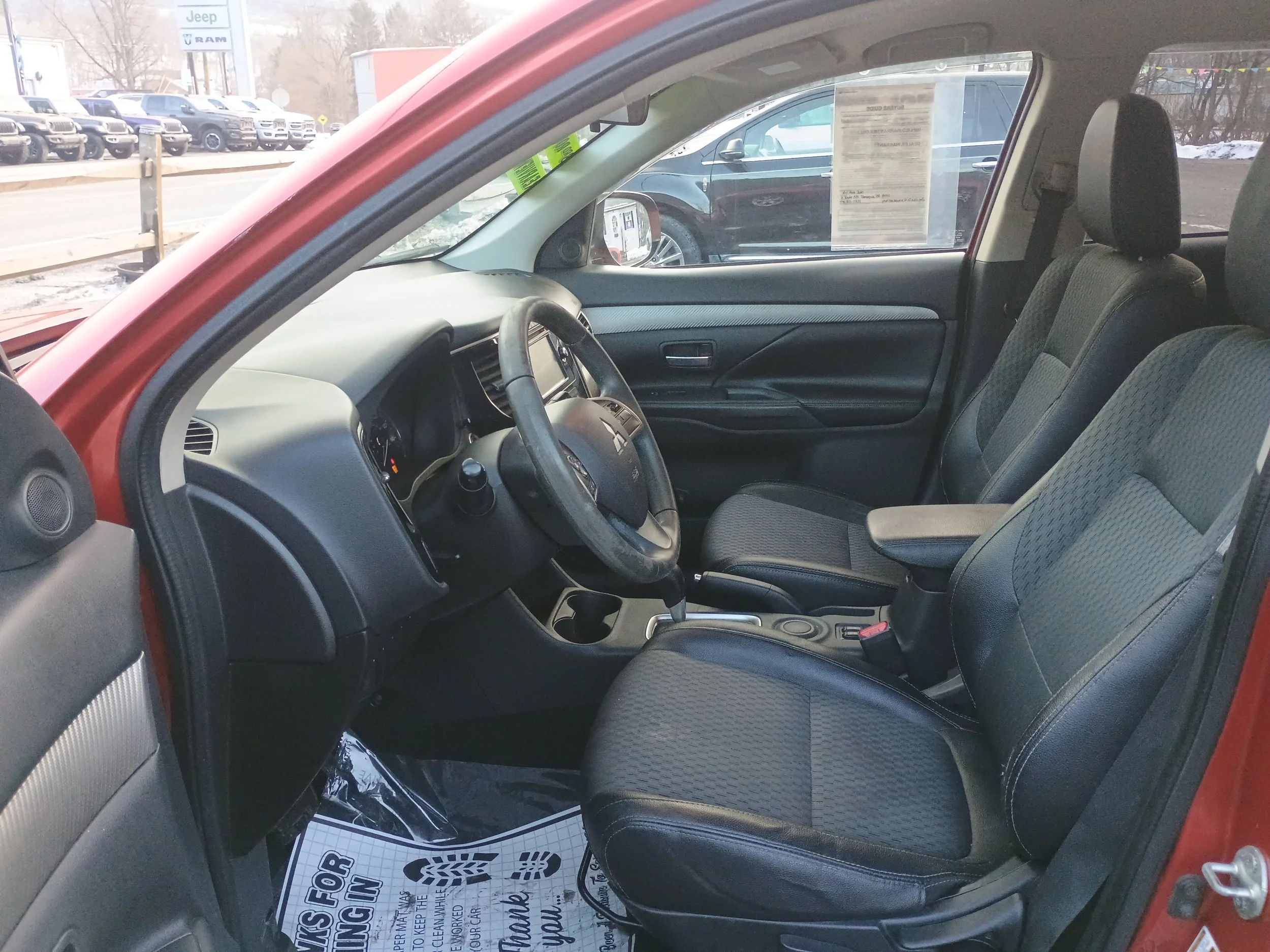 Interior of a red car showing black fabric seats, steering wheel, dashboard, and driver side door, parked in a lot with other vehicles visible outside.