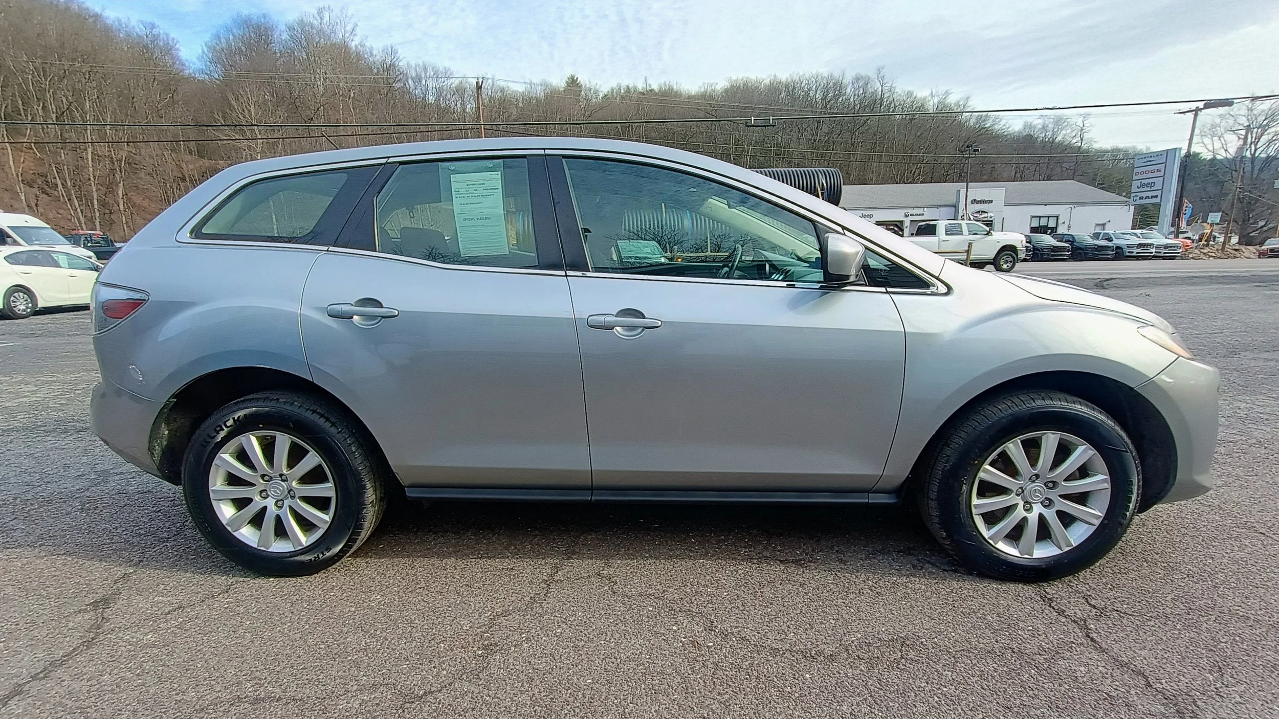Side view of a silver crossover SUV parked on a lot at a car dealership with other vehicles and a dealership building in the background.