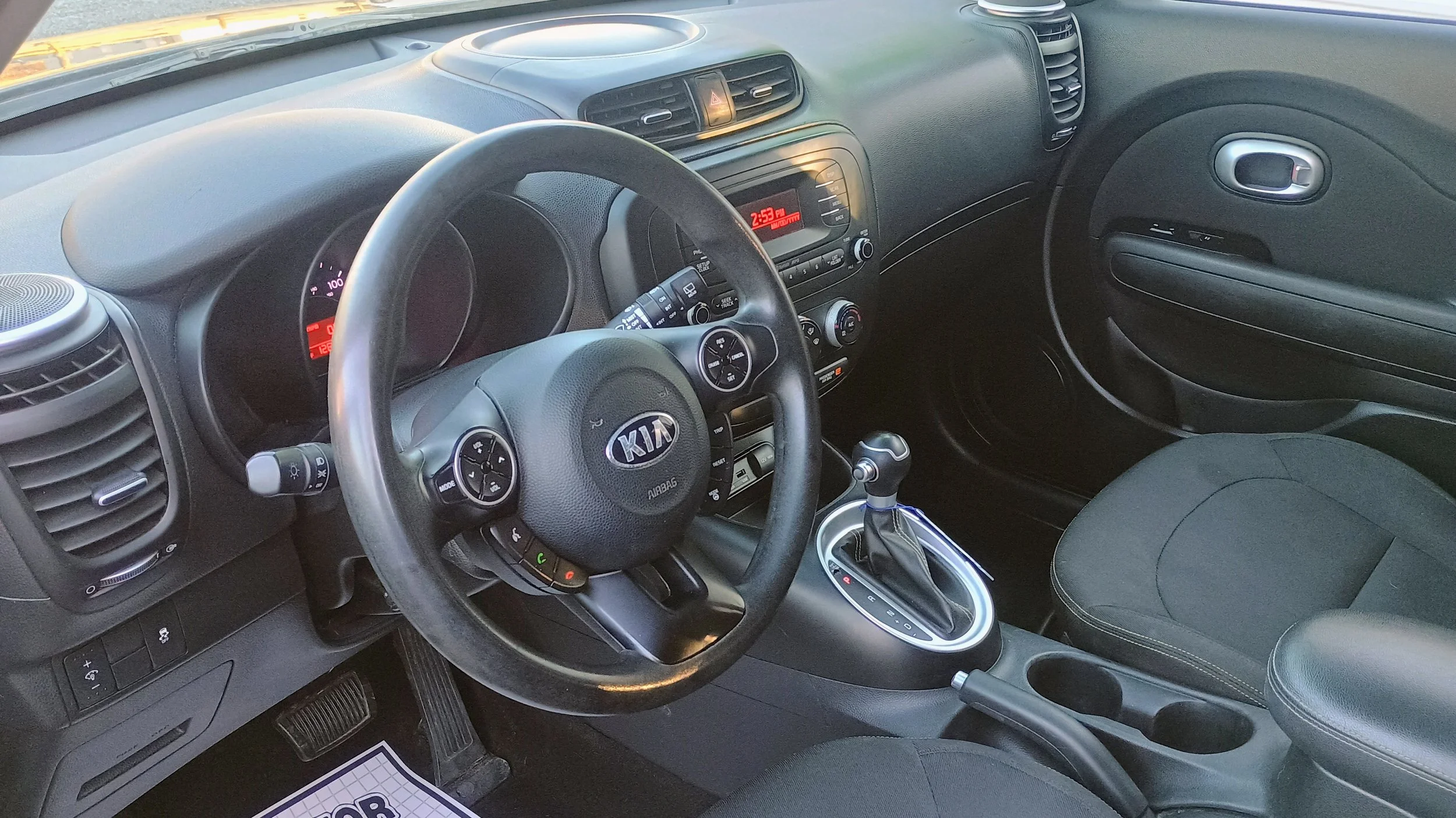 Interior of a Kia car showing the steering wheel, dashboard with digital clock, gear shift, and front passenger seat.