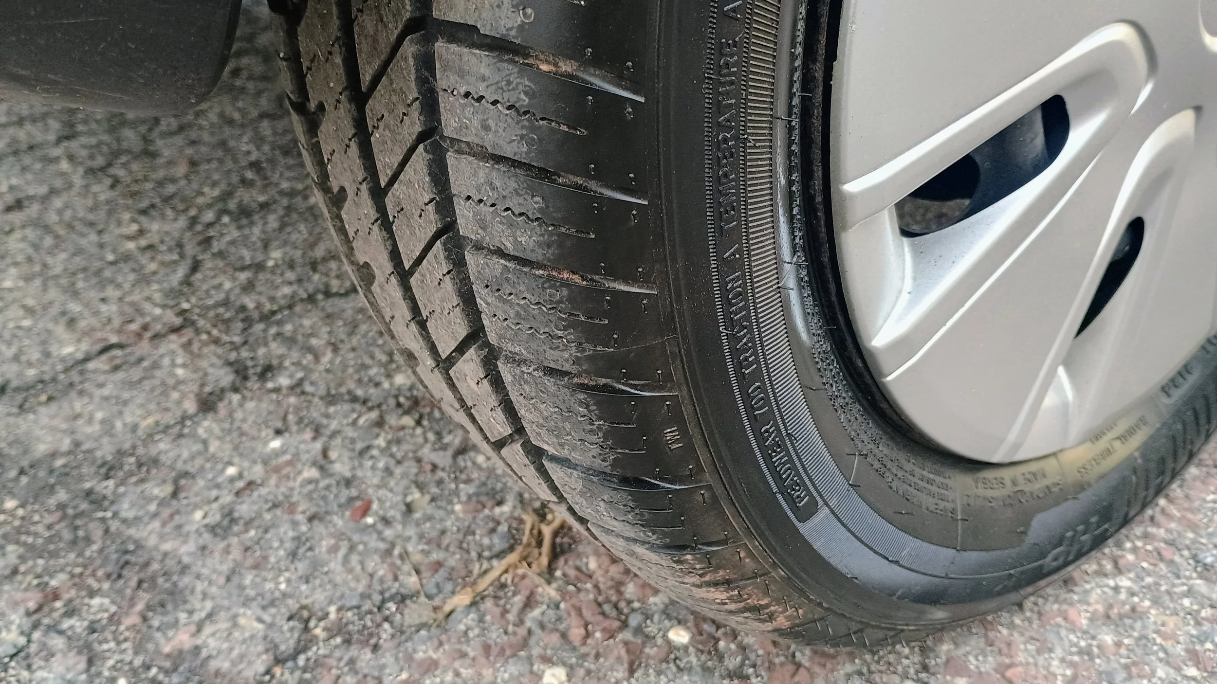 Close-up of a car tire with tread patterns, mounted on a silver alloy wheel, on a gravel surface.