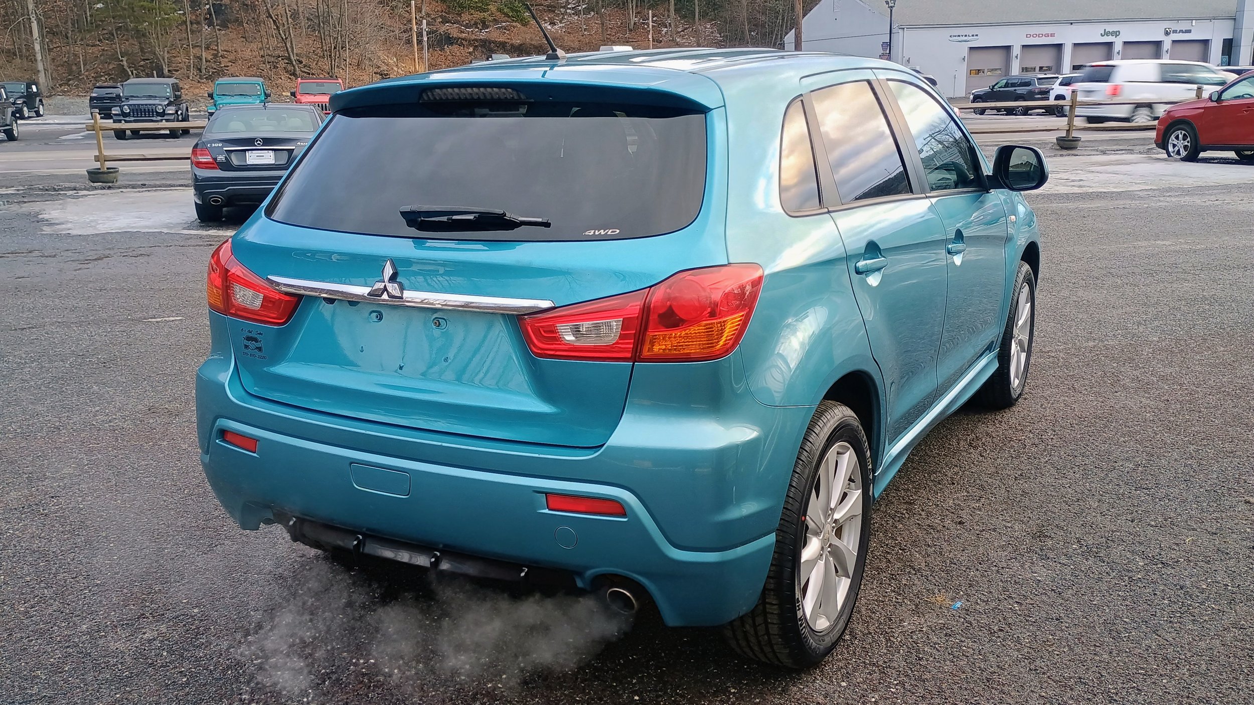 A blue Mitsubishi hatchback car parked in a lot with other vehicles, some of which are blurred from motion, in the background. The car is facing away from the camera, showing its rear and passenger side.