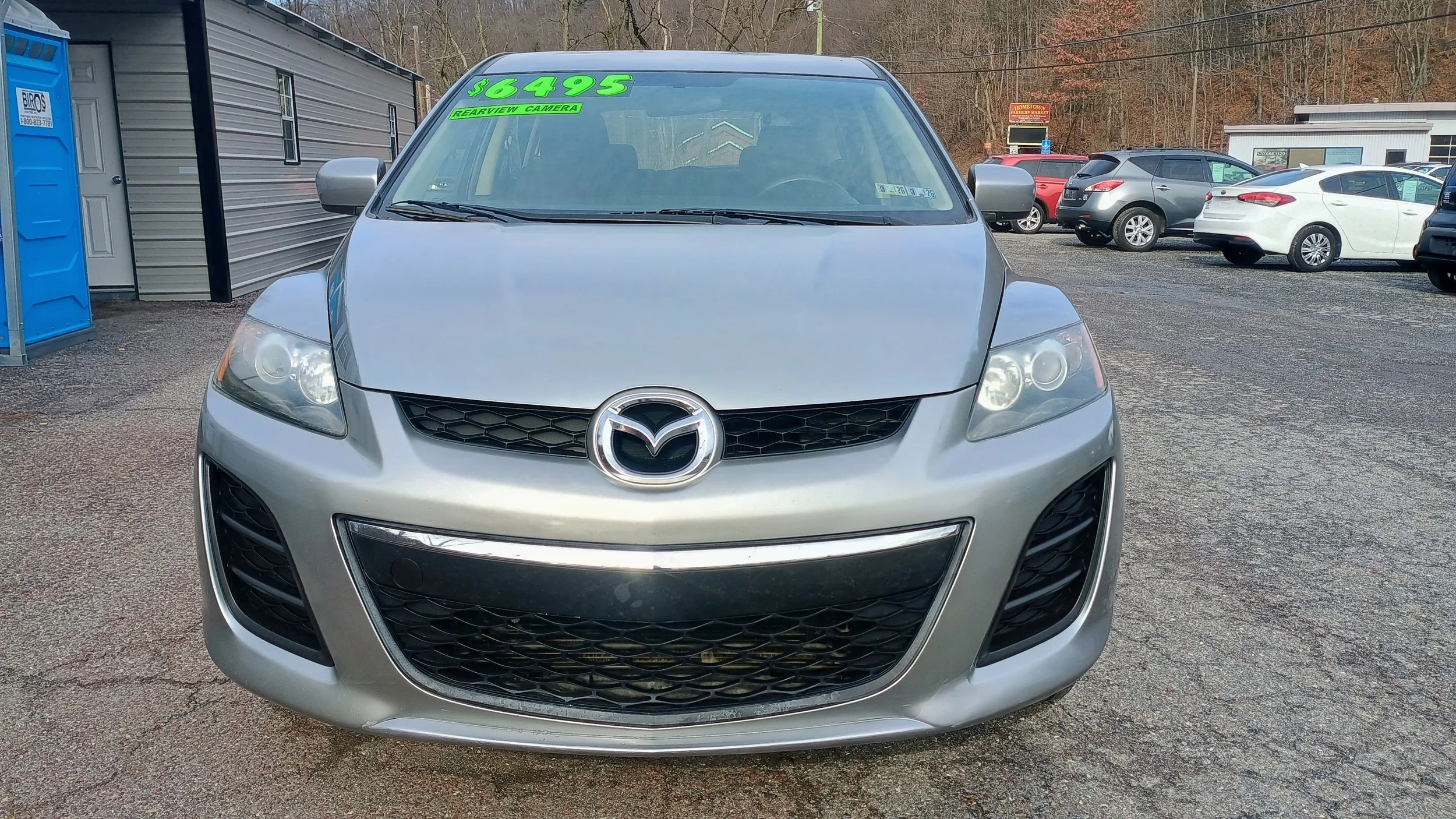 Front view of a silver Mazda car with a green price and feature sticker on the windshield, parked at a used car lot with other cars in the background.