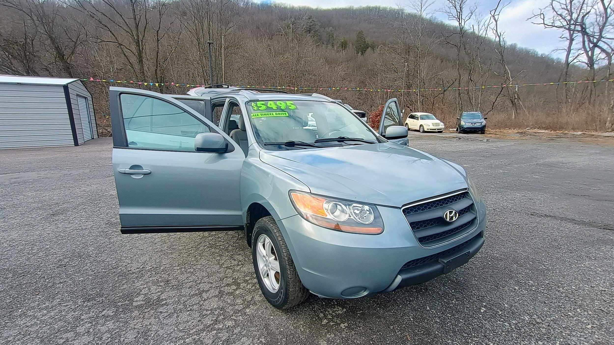 Silver Hyundai SUV parked on a gravel lot with its front and driver's side doors open, displaying a bright green sign on the windshield that reads '5495 ALL WHEEL DRIVE.' In the background, there are a few other cars, leafless trees, a hillside, and a small storage shed.