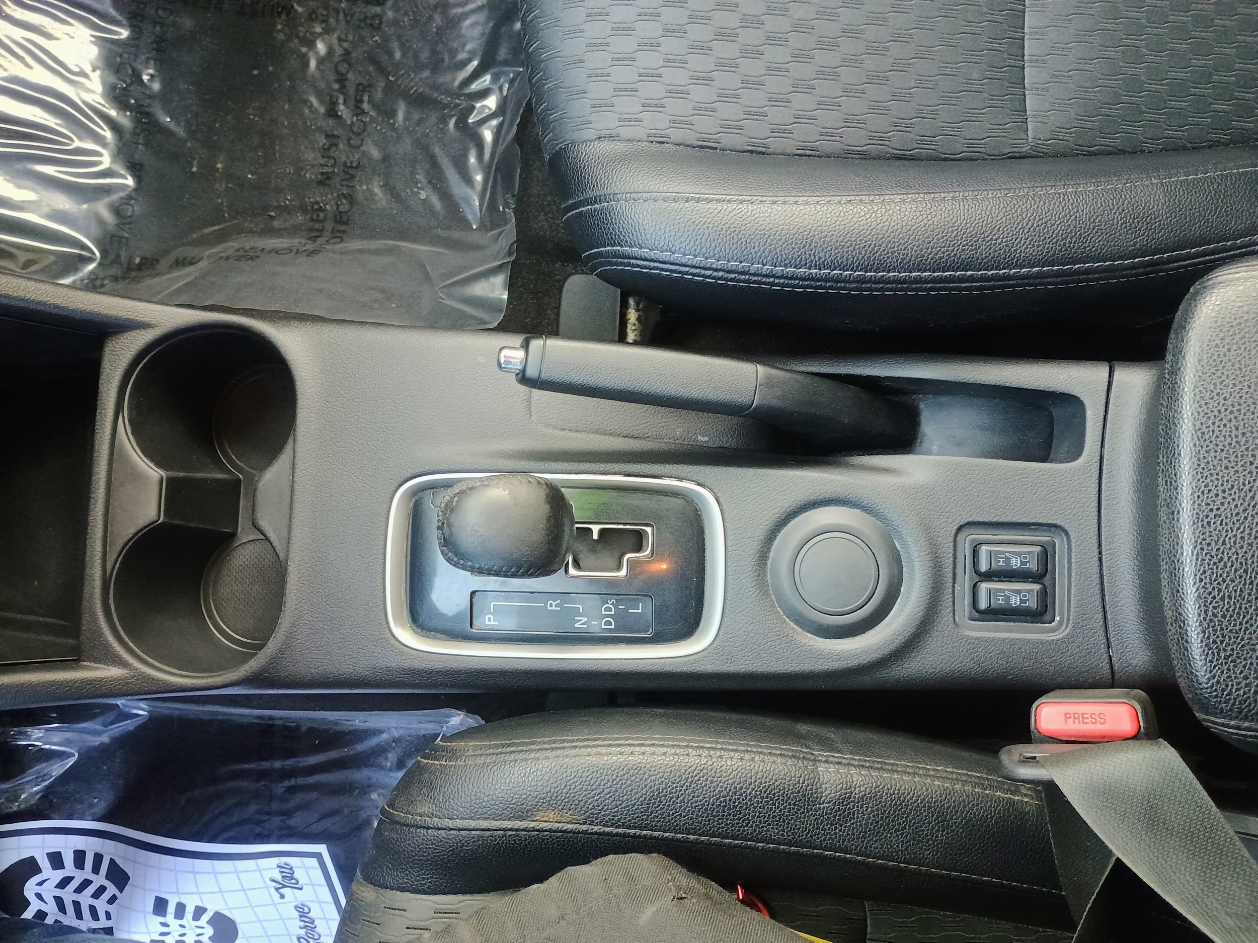 View of a car's center console, showing an automatic gear shift, handbrake, two cup holders, a circular control button, and buttons for heated seats. Part of the driver's seat and seatbelt are also visible.