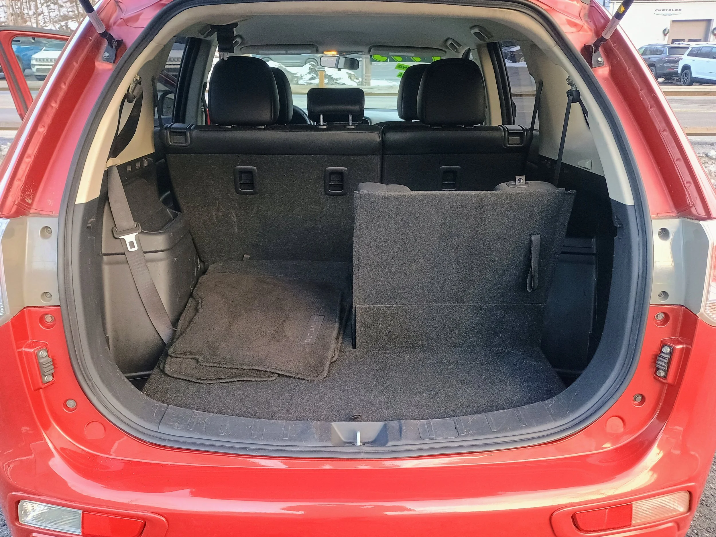 The open cargo area of a red SUV showing the folded-down rear seats, a black floor mat on the left side, and a partially upright rear seat on the right, with parked vehicles visible in the background.