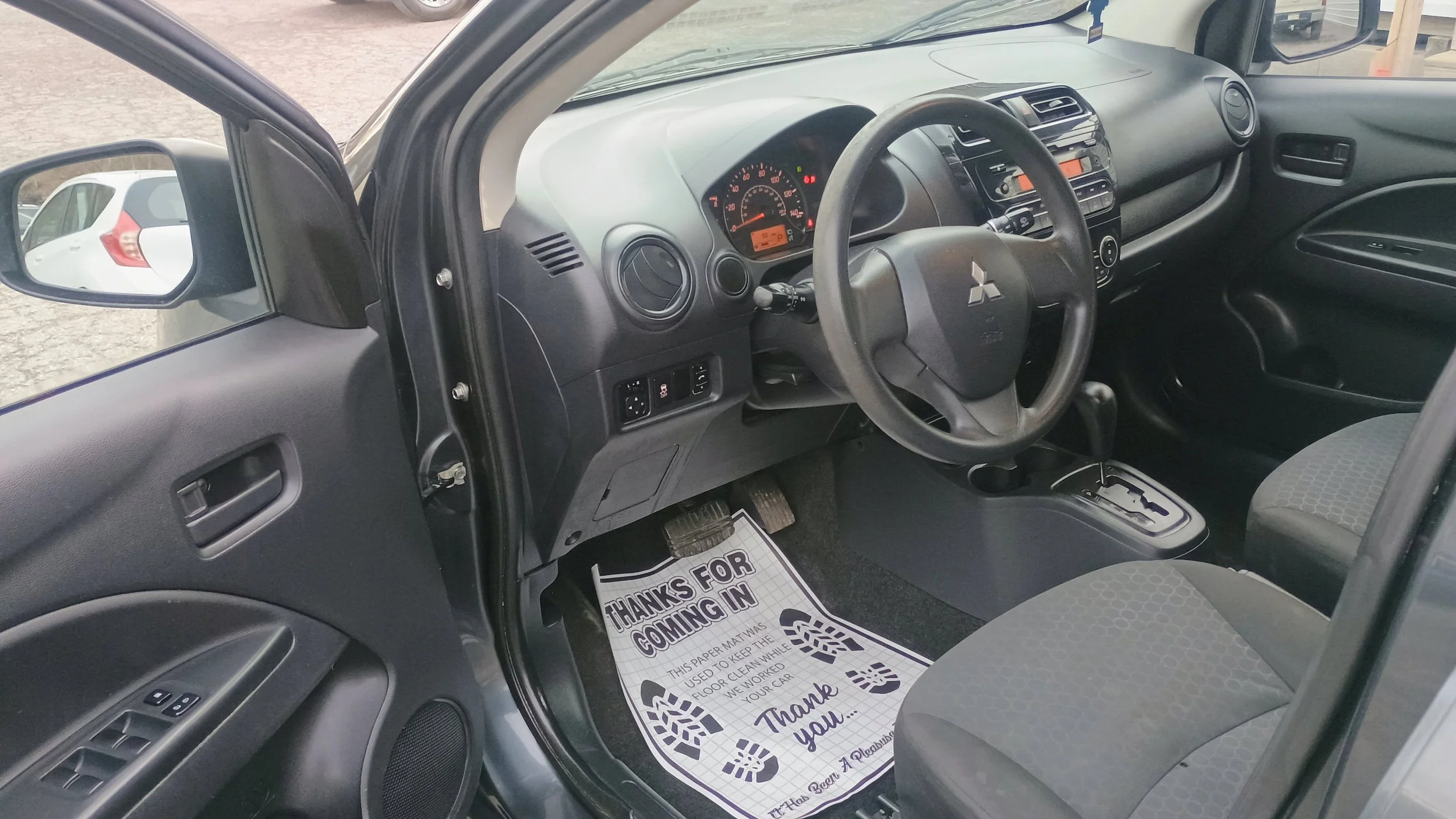 Interior view of a Mitsubishi car showing the dashboard, steering wheel, gear shift, and front seats, with a paper floor mat that says "Thanks for coming in."