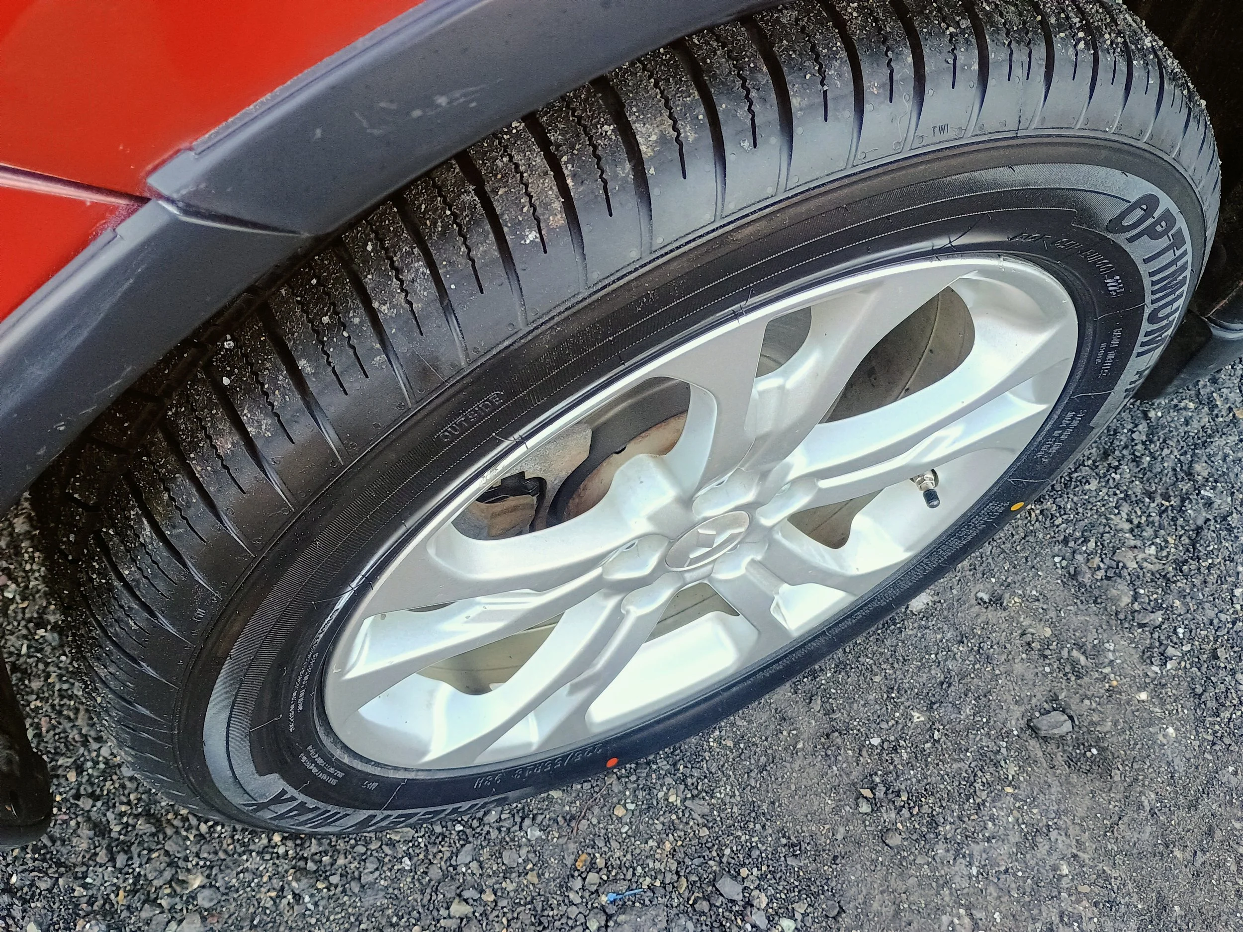 Close-up of a car tire mounted on a white rim, with black tread and sidewall, parked on gravel ground.