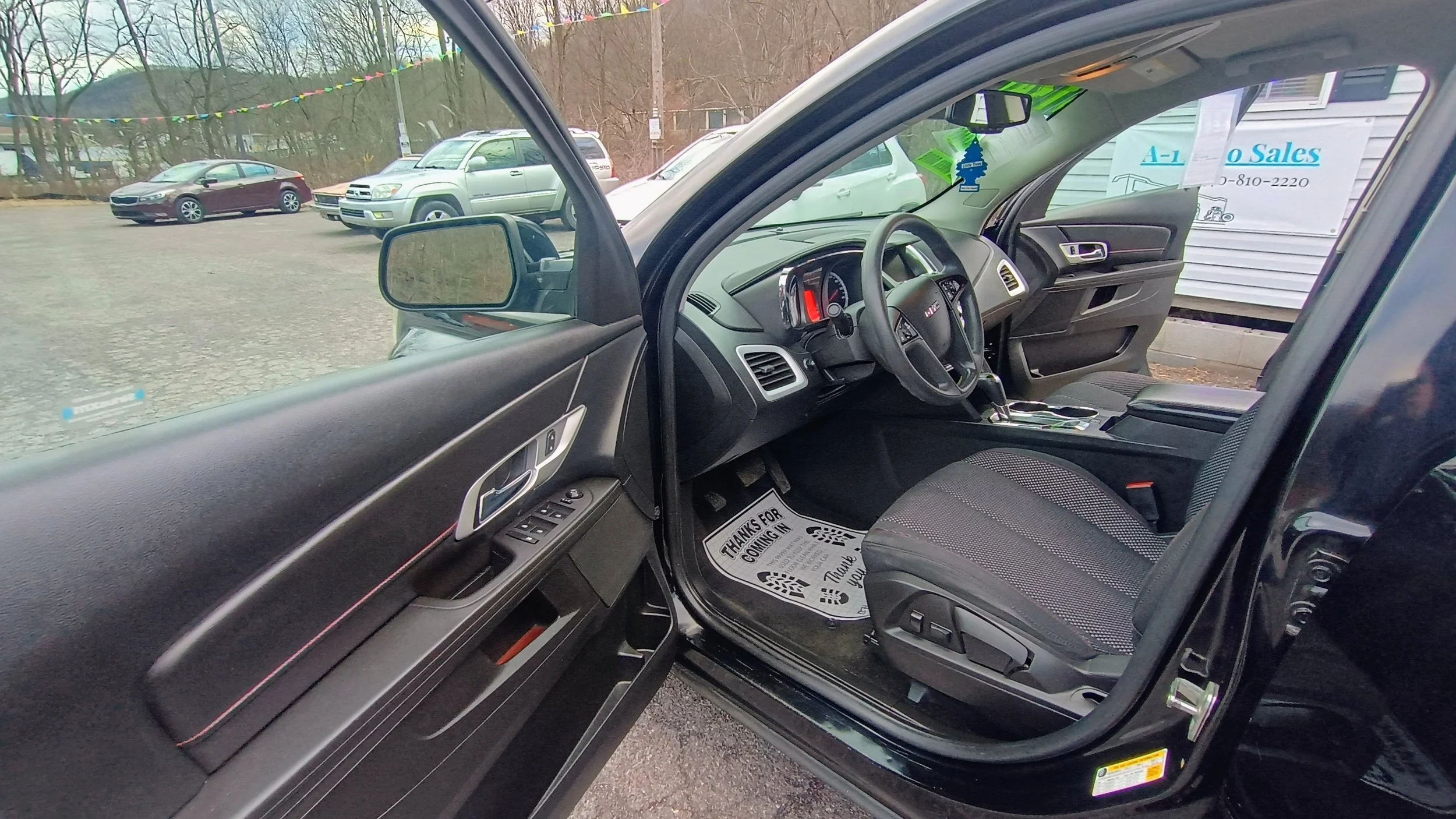 Inside the front passenger side of a black car, showing the dashboard, steering wheel, and front seat, with other vehicles in a parking lot outside.