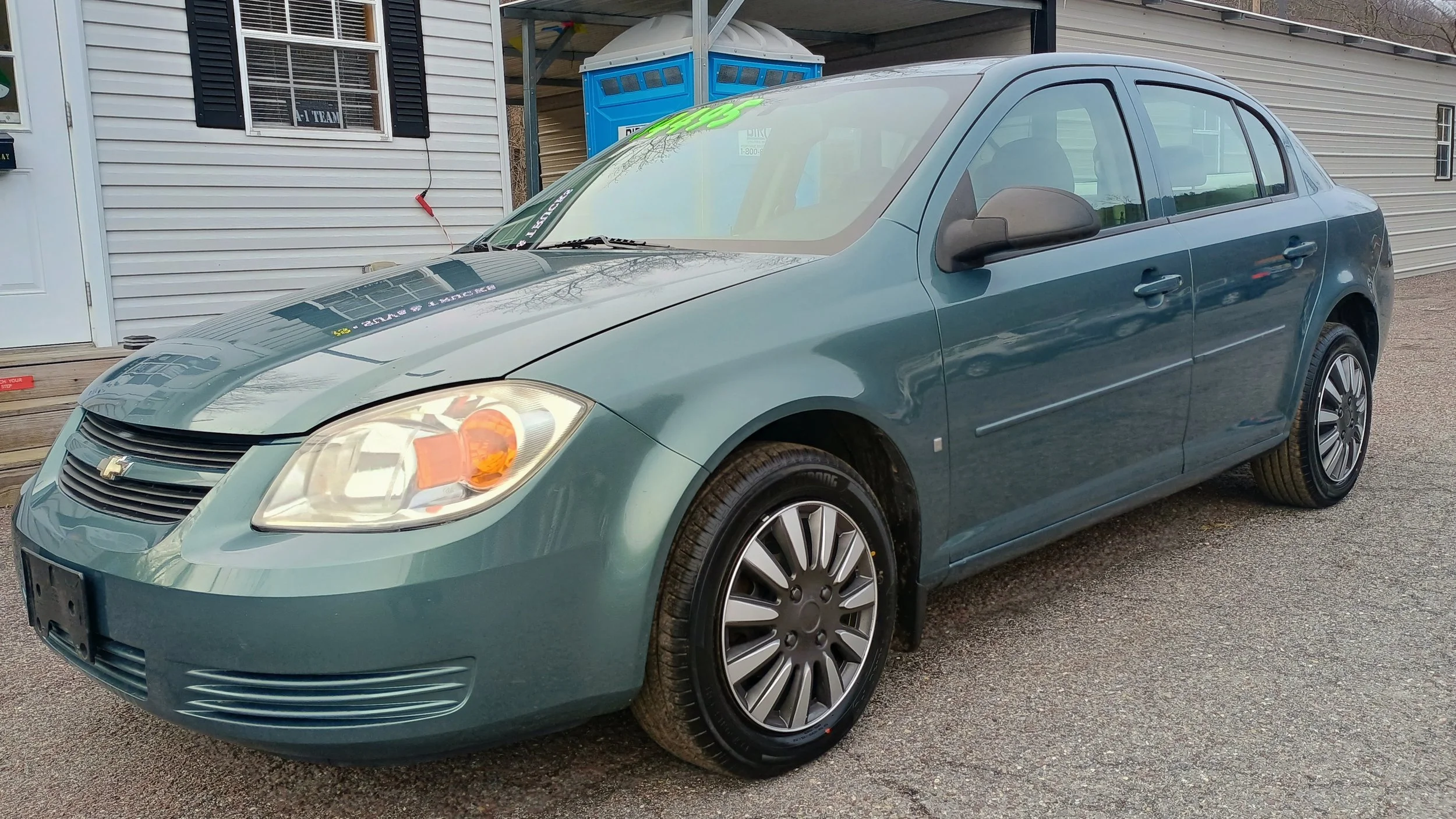 A teal-colored four-door sedan parked on a gravel surface in front of a mobile home with a white exterior, black shutters, and a window. A blue portable toilet is visible in the background.