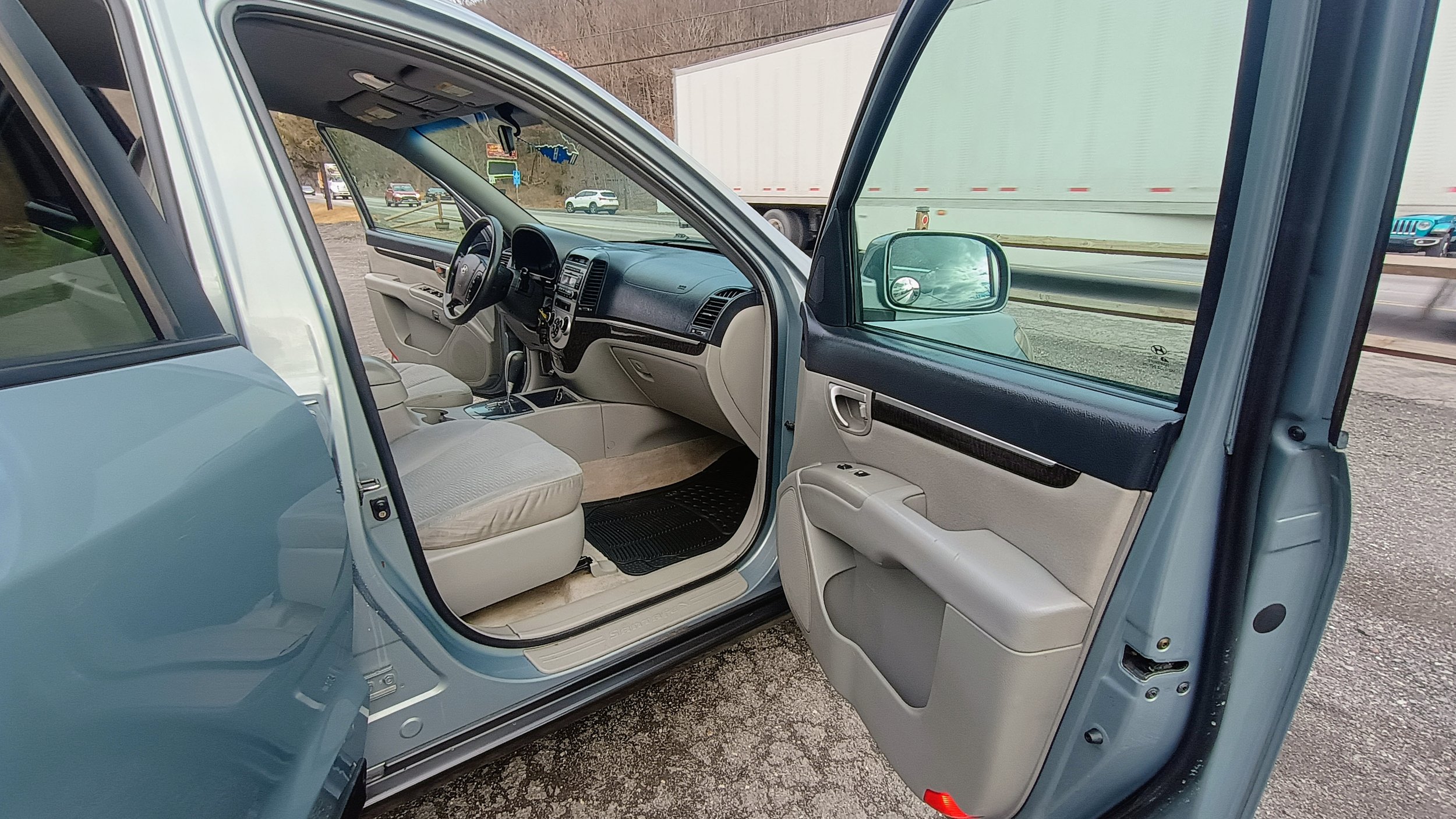 The image shows the open front passenger door of a light blue vehicle, revealing the beige interior and dashboard with the steering wheel and gear shift. Outside, part of a highway with a large white truck and some cars in the background.