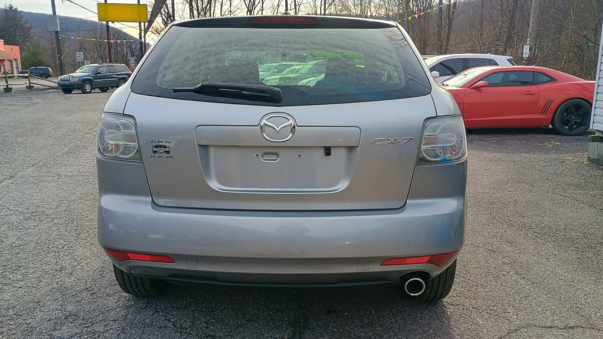 Rear view of a silver Mazda CX-7 parked on a gravel lot with other vehicles and trees in the background.