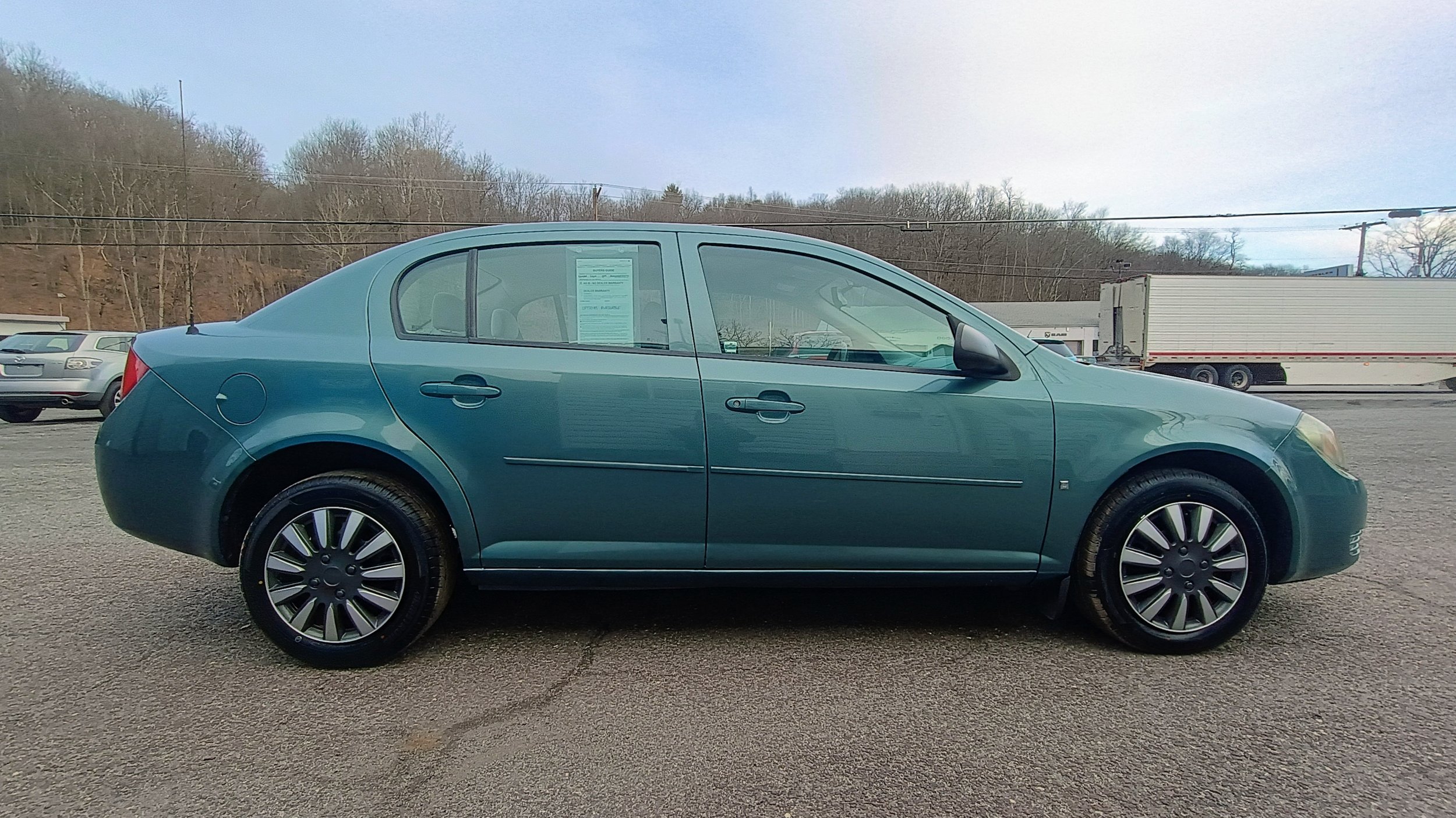 Side view of a teal sedan car parked on a gravel lot with other cars and trucks in the background.